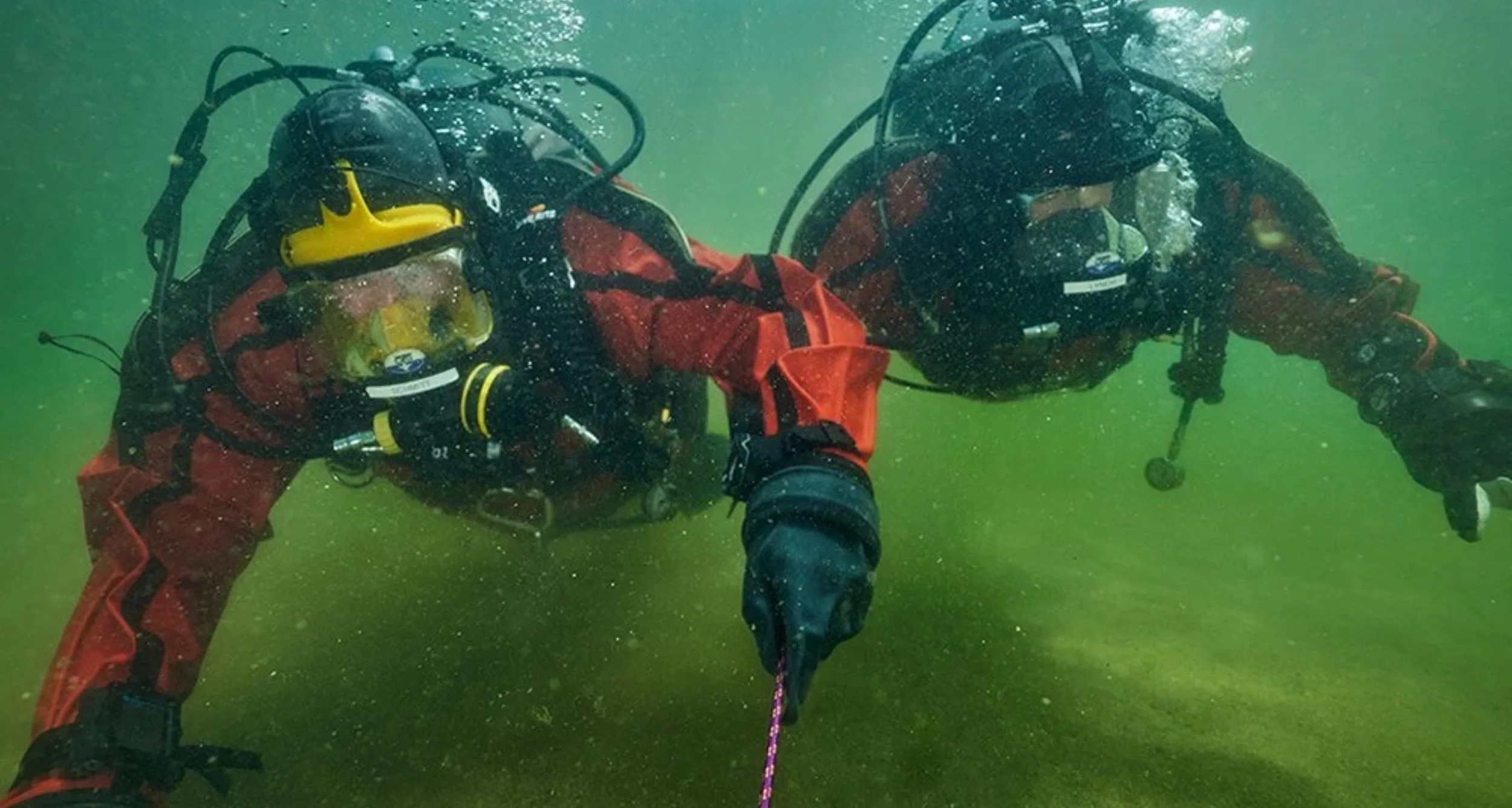 Two scuba divers in red suits underwater, holding hands, with one holding a rope, surrounded by green water.