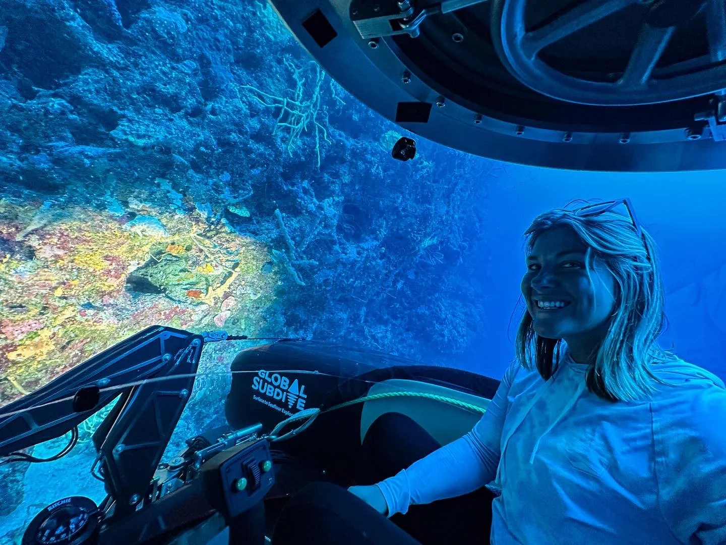 A woman smiling inside a submarine looking out at a vibrant coral reef underwater.