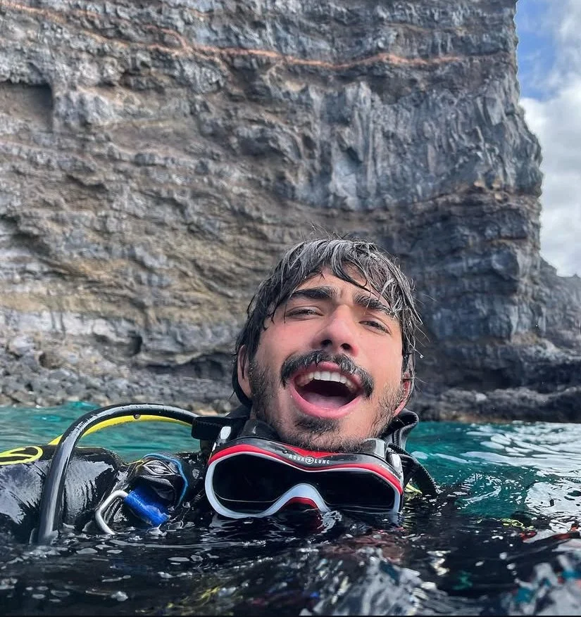 A man with wet hair and a beard, smiling and singing or shouting, is floating in the water near a rocky cliff, holding scuba diving gear.