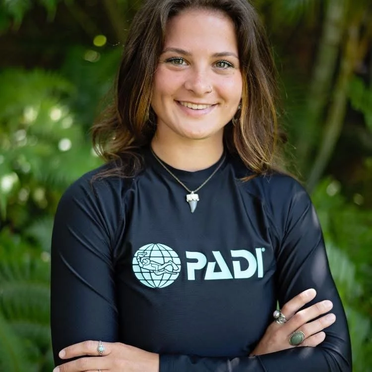 Young woman smiling outdoors wearing a black PADI diving shirt with her arms crossed, surrounded by green foliage.