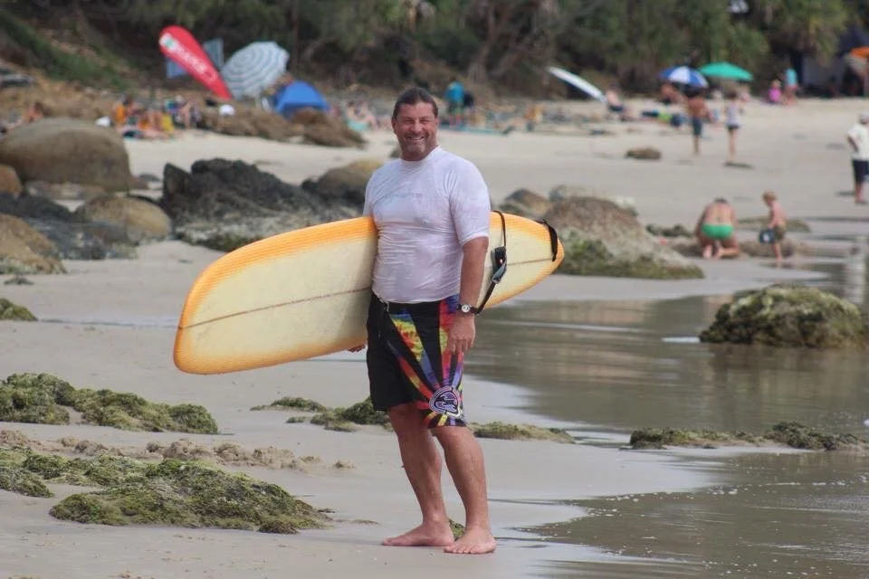 A man smiling on a sandy beach holding a yellow surfboard, with people and umbrellas in the background.