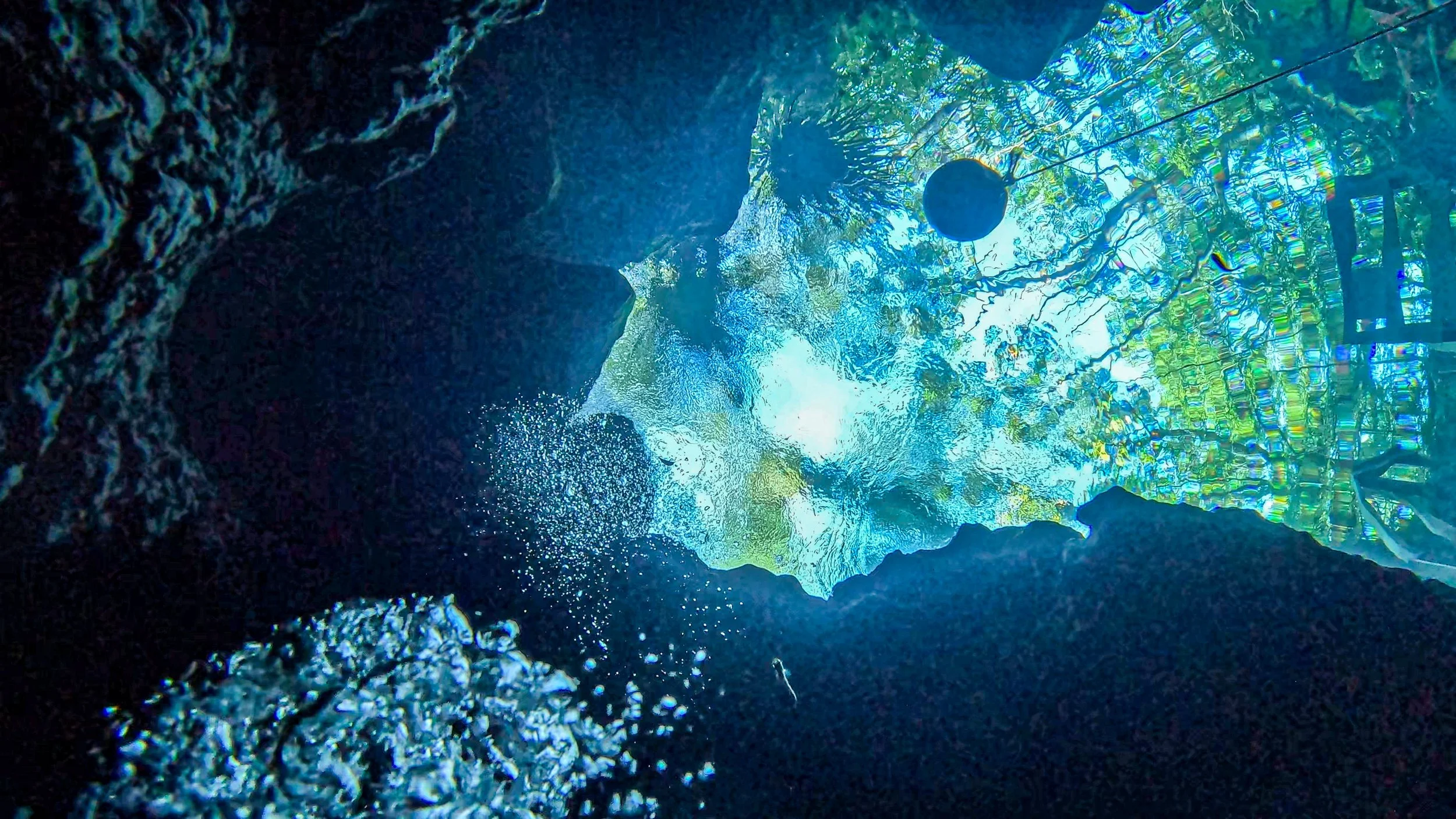 View from inside a cave looking upward toward sunlight, with rocks and water droplets surrounding the opening, and trees visible outside.