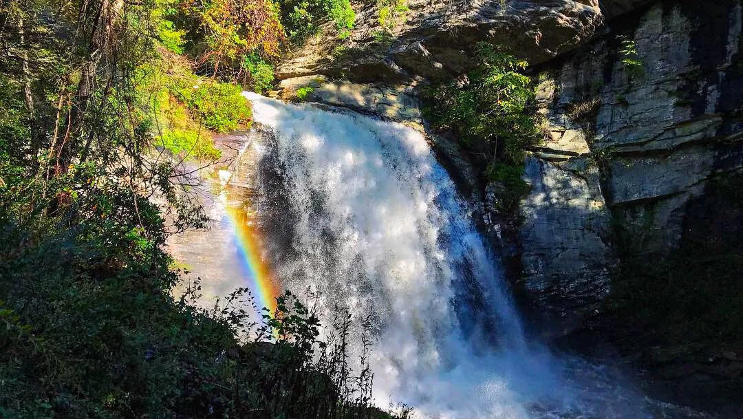 A waterfall cascading down a rocky cliff into a river below, with a rainbow visible in the mist, surrounded by green trees and foliage.