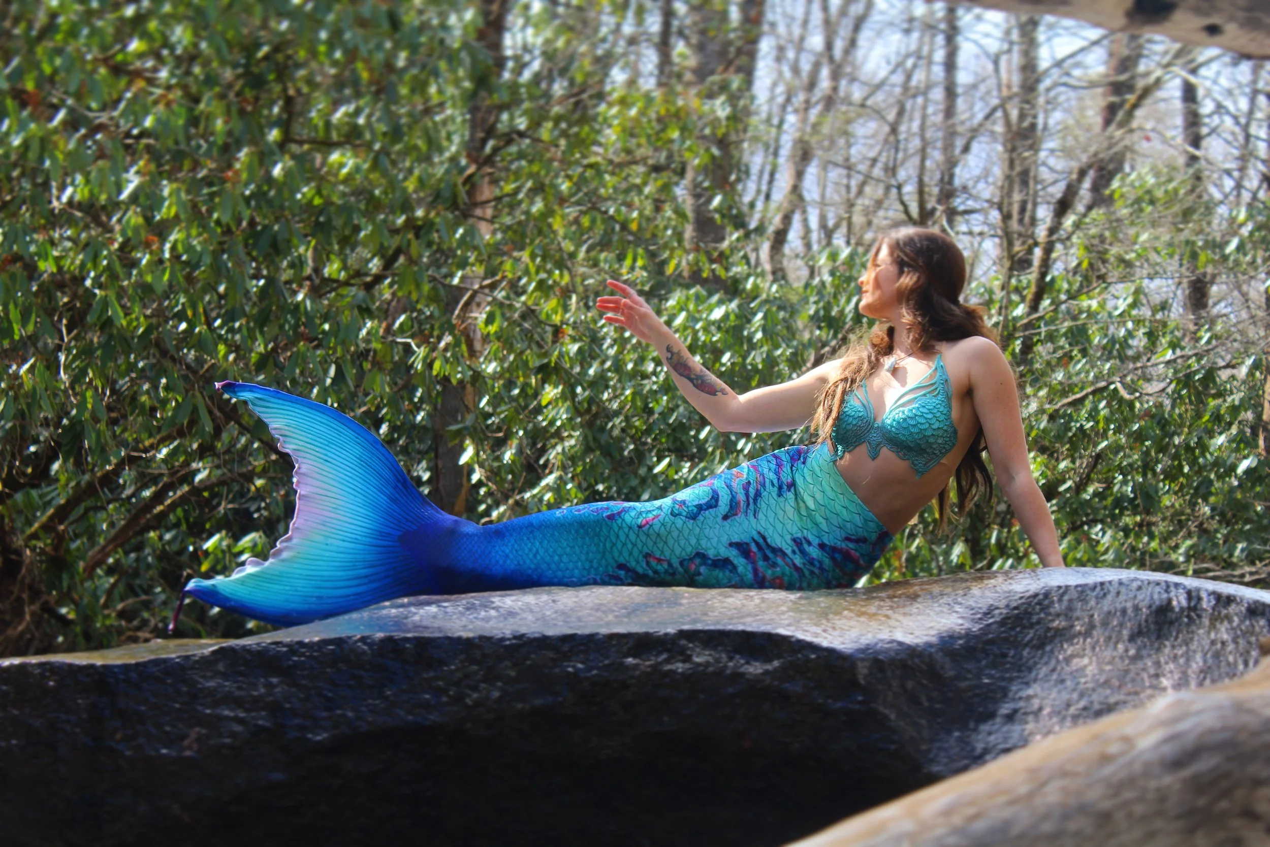 A woman dressed as a mermaid with a teal top and blue tail, lying on a rock in a wooded outdoor setting.
