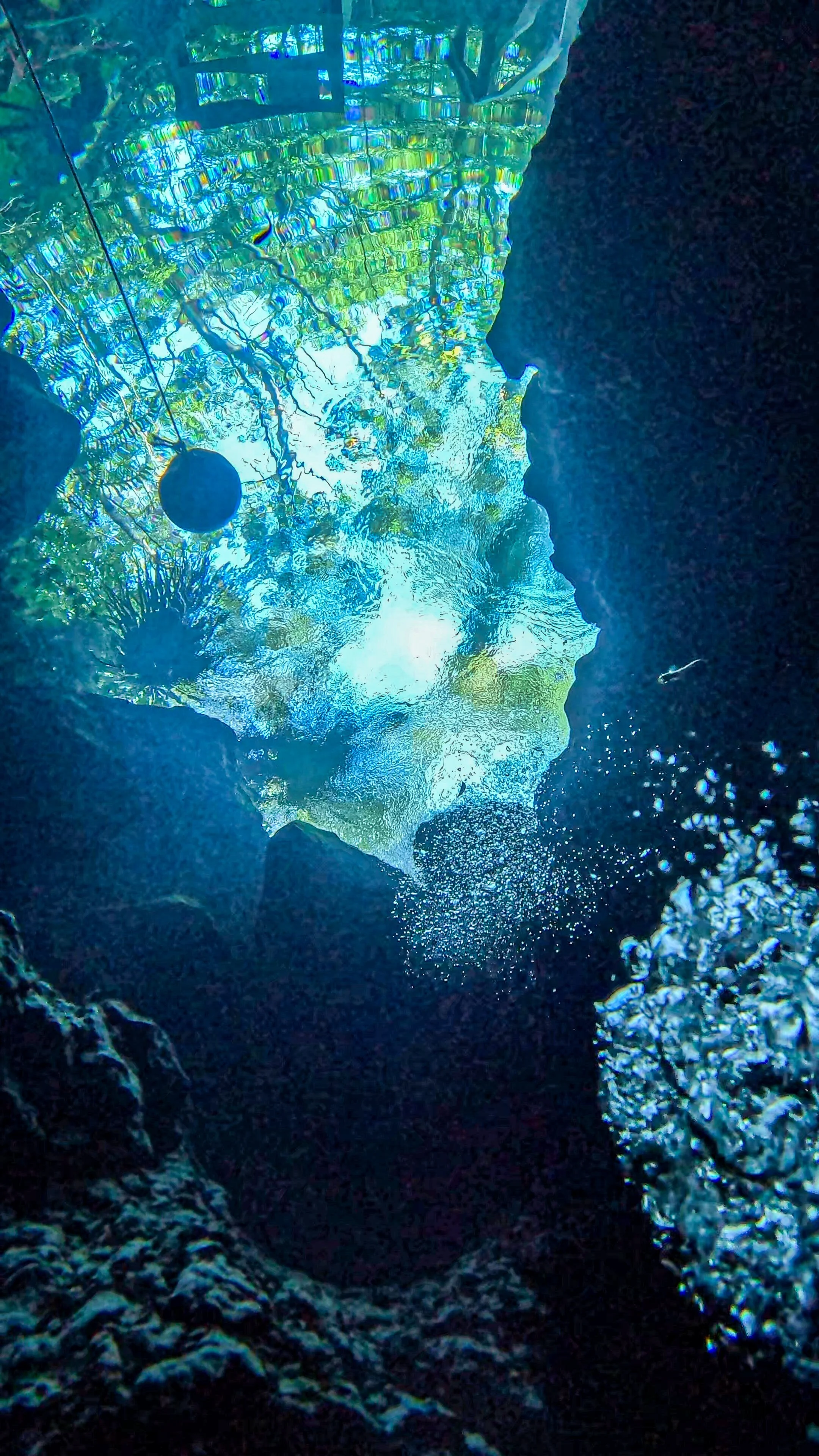 Underwater view of a rock formation with bubbles rising to the surface, showing sunlight filtering through the water and rocks.