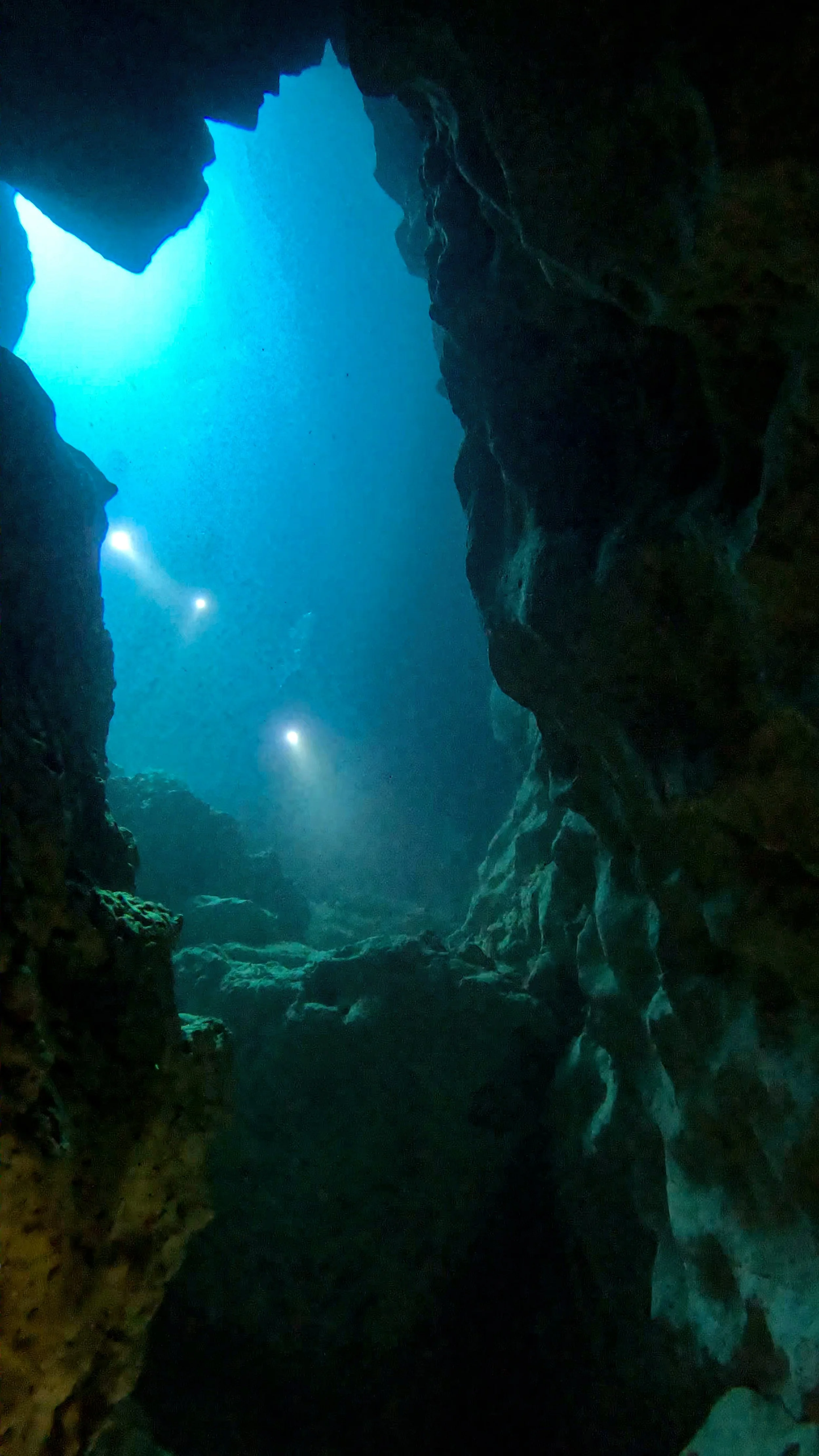 Inside a dark underwater cave with rocky walls, illuminated by blue sunlight coming from above and a few small underwater lights.