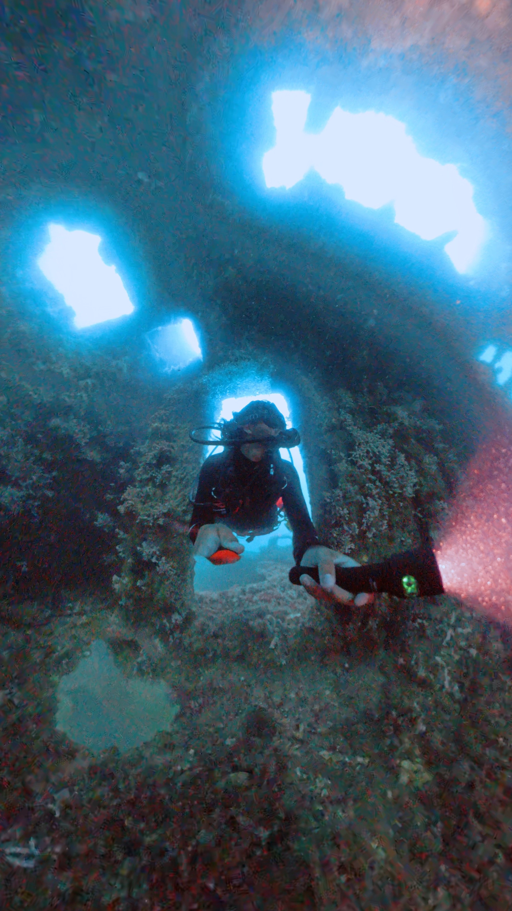 A scuba diver underwater, taking a selfie inside a shipwreck or underwater structure with bright blue light filtering through openings above.