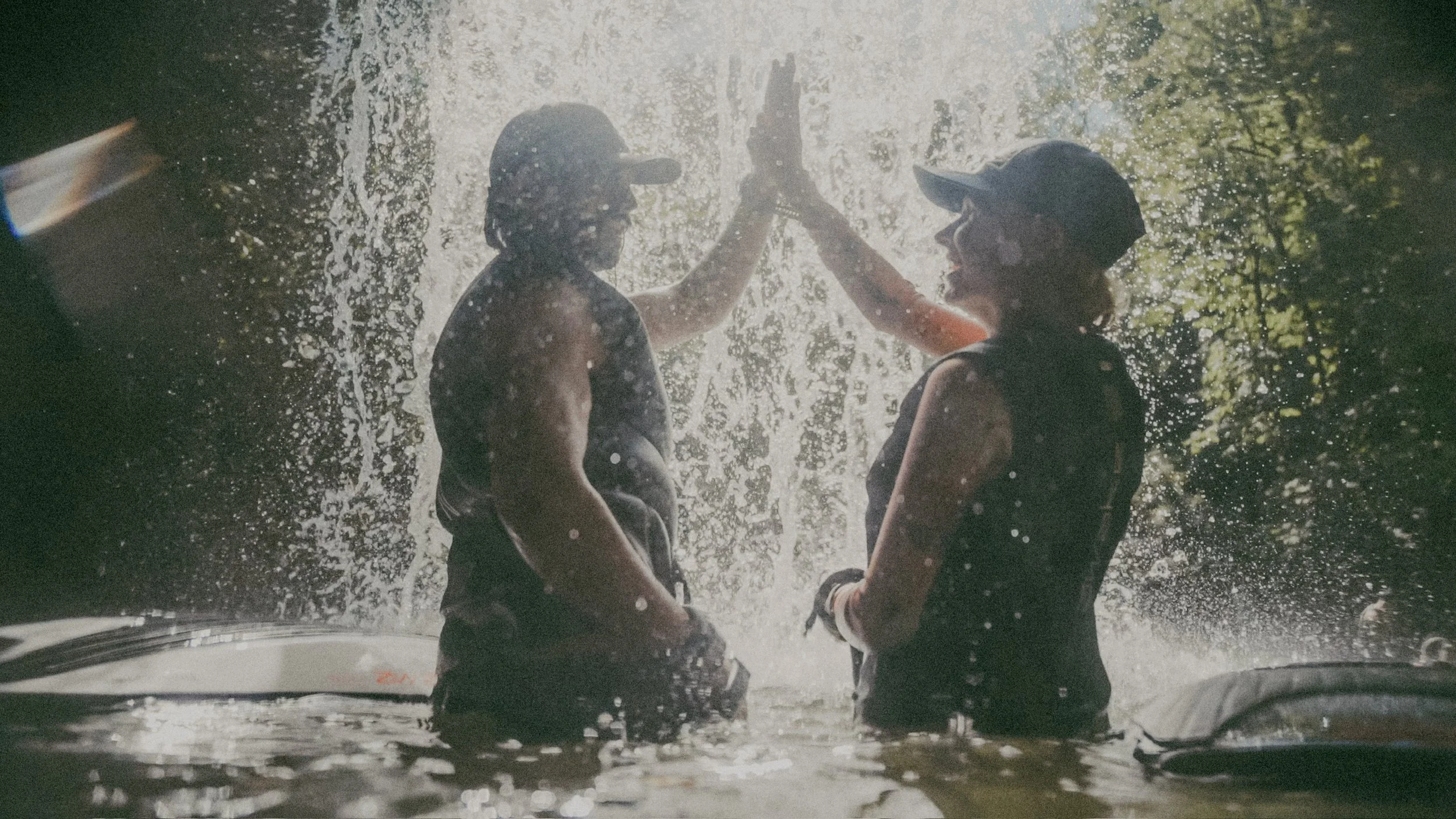 Two people, a man and a woman, wearing caps and high-visibility vests, are high-fiving each other underwater during a waterfall or rain. Bright sunlight filters through trees in the background.