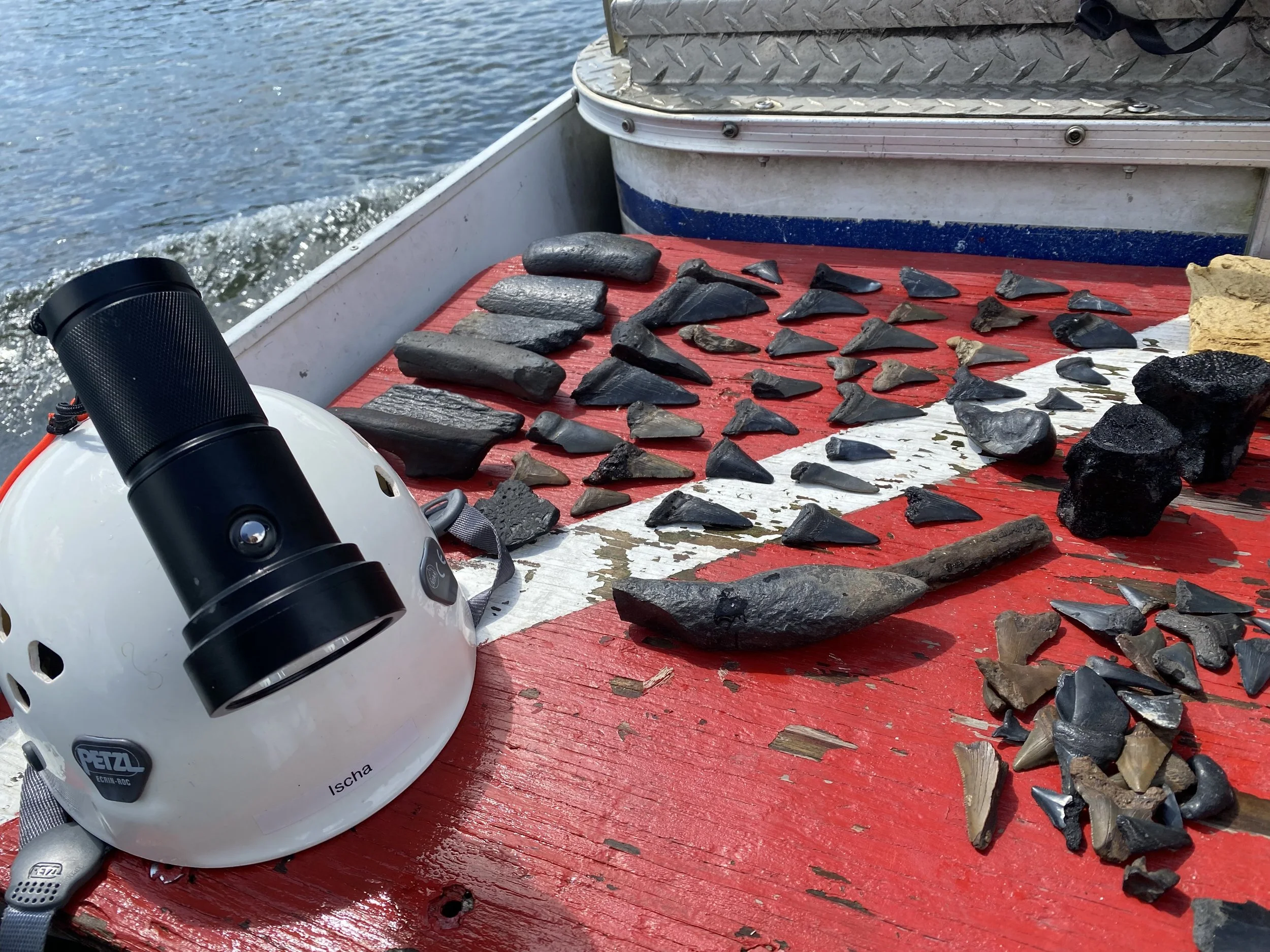 Row of assorted arrowheads and stones on a red and white boat surface, with a black flashlight and a white helmet nearby, part of a boat water scene.