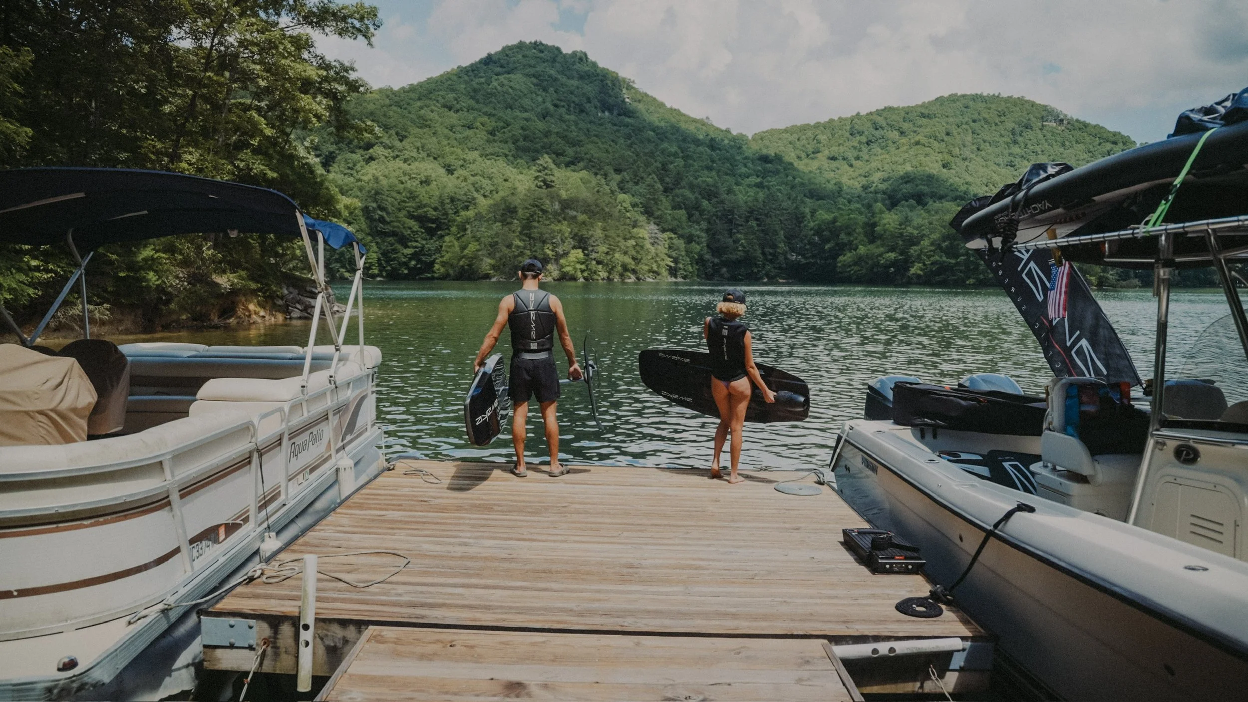 Two people standing on a wooden dock by a lake, holding paddleboards, with boats on either side and green hills in the background.