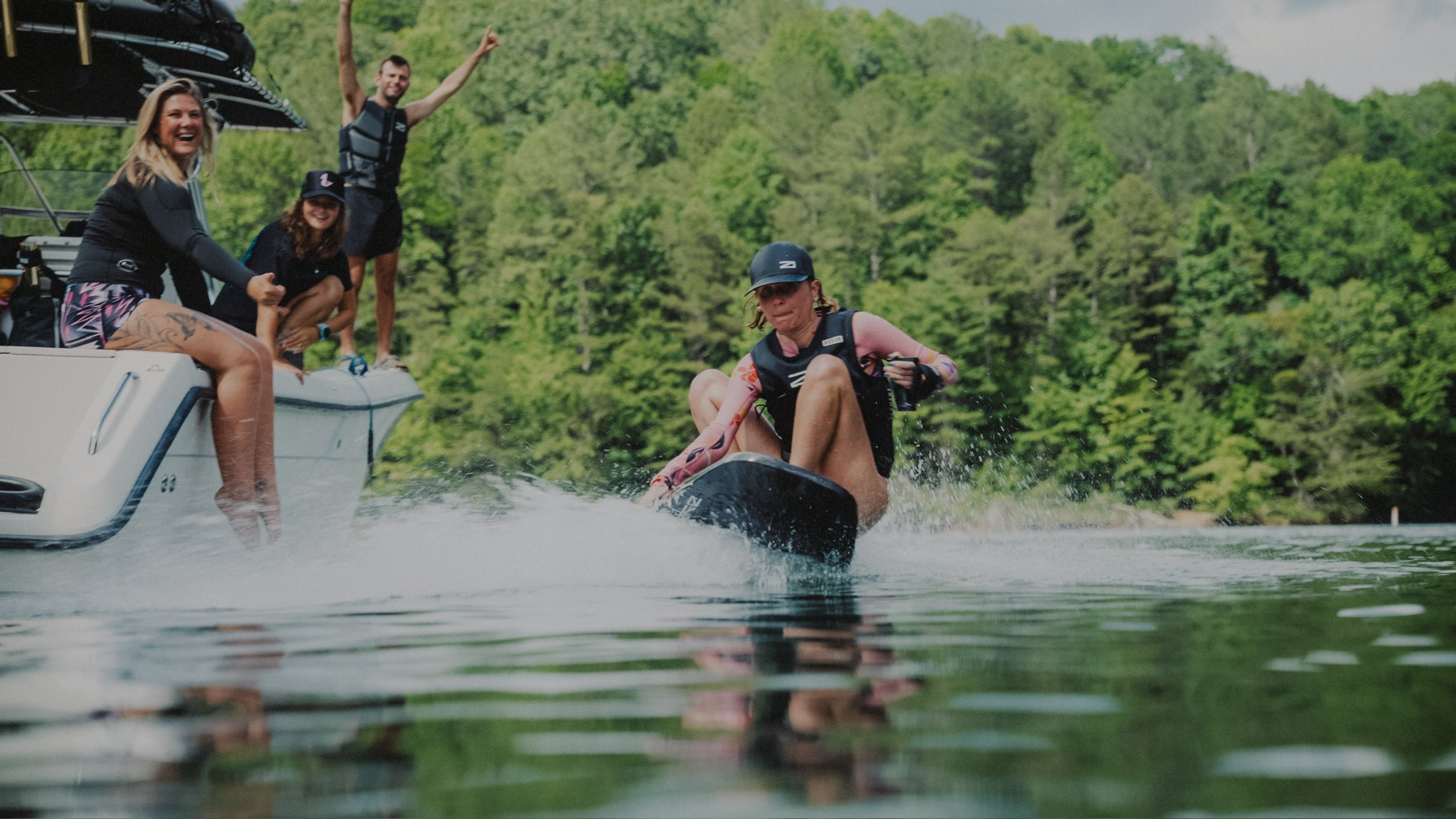 Group of people on a boat and a person wakeboarding on a lake surrounded by green trees.