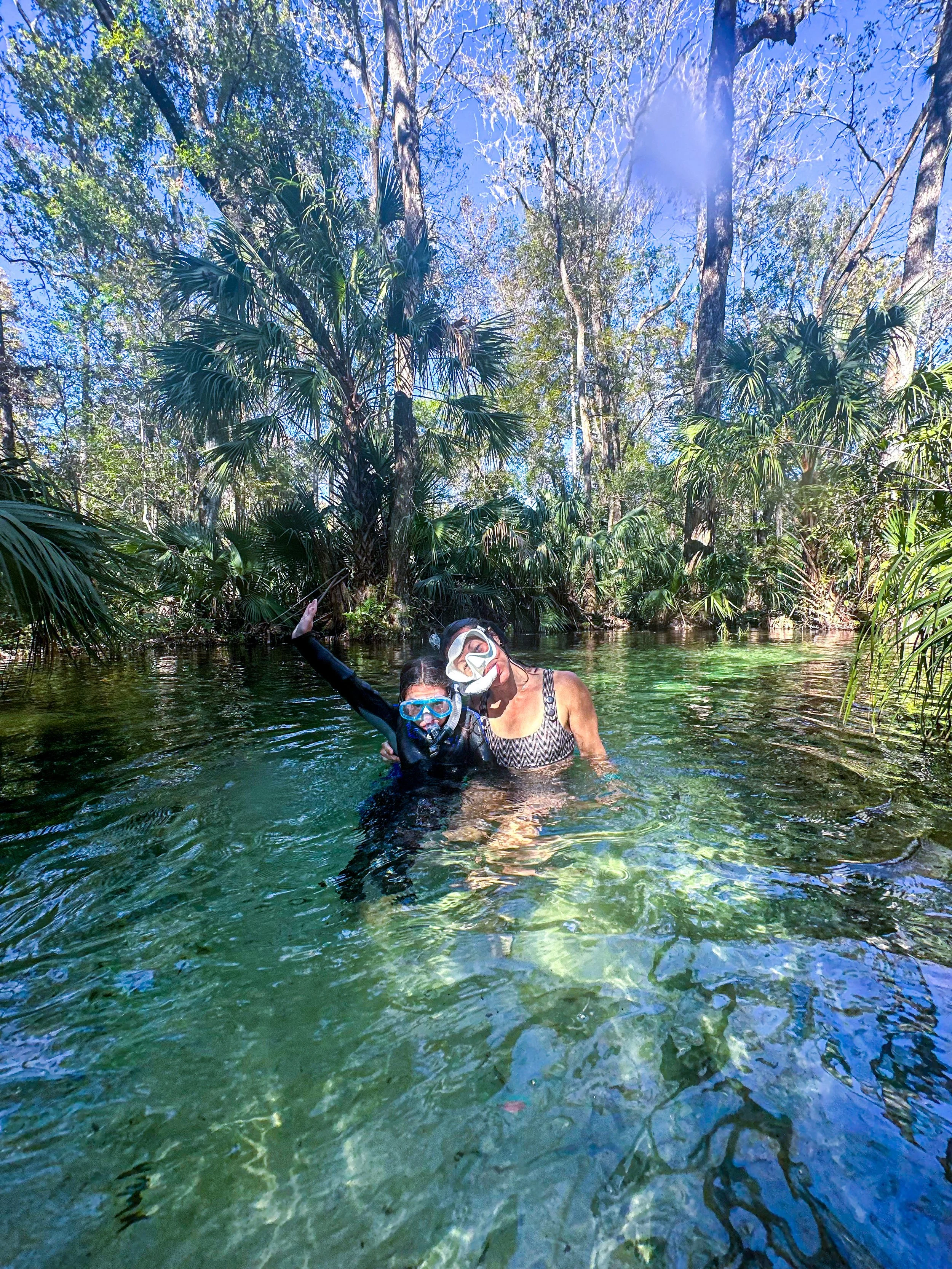 Two people wearing snorkeling masks and wetsuits, smiling and waving in a clear water creek surrounded by lush green trees and vegetation.