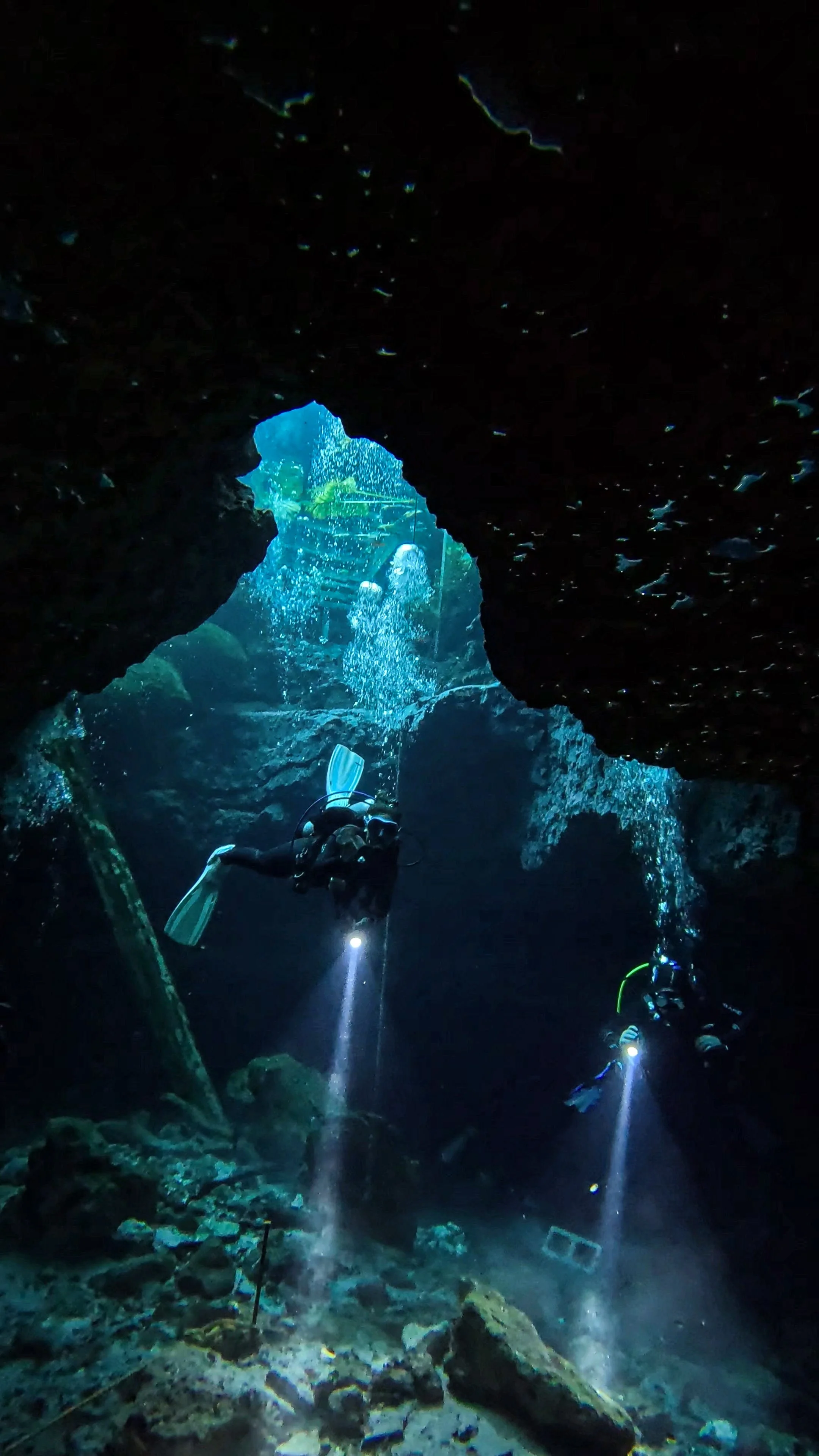 Two scuba divers exploring an underwater cave with rocks and boulders, holding flashlights that illuminate the seabed.