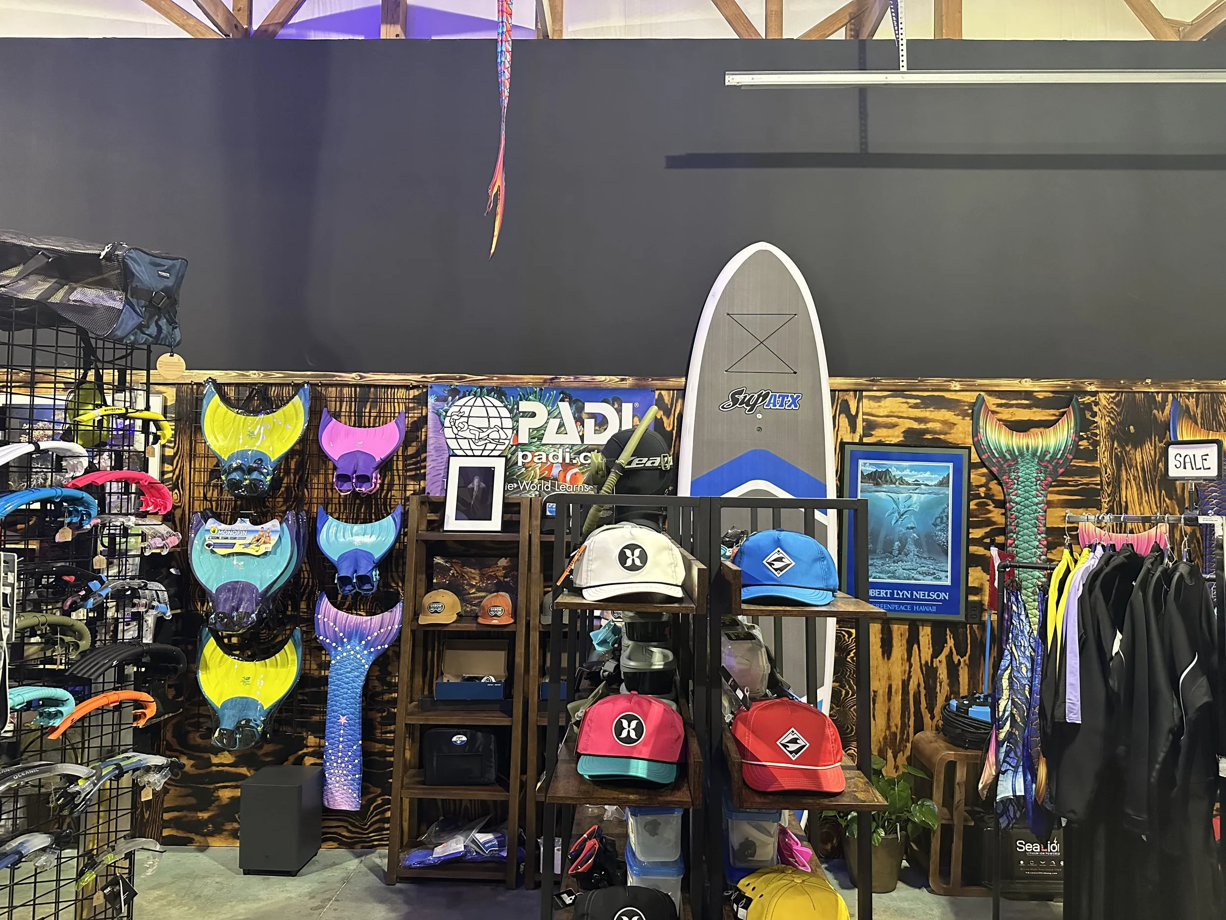 Display of surfing gear including fins, hats, and accessories, with a surfboard, framed ocean photograph, and sale sign in the background.