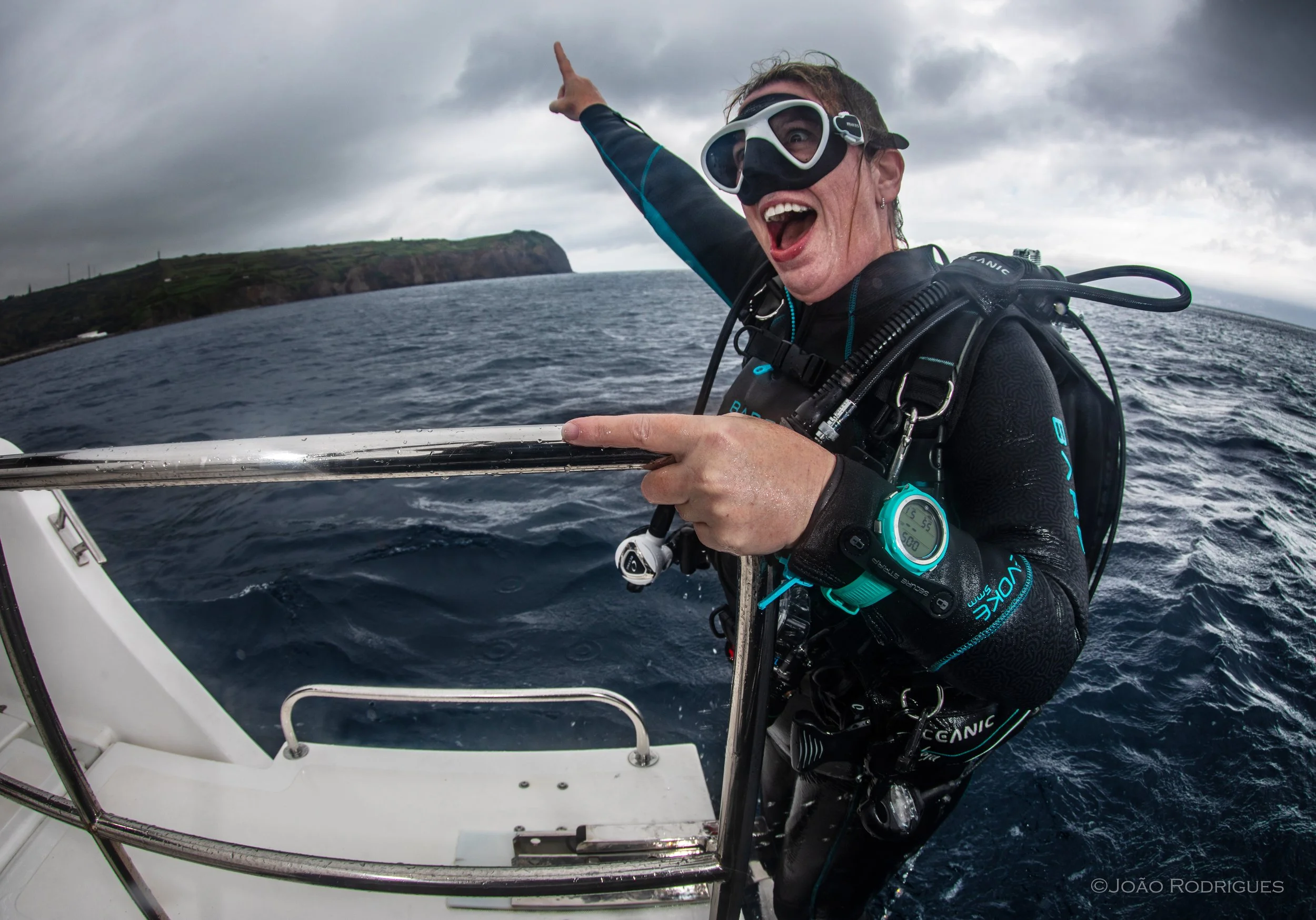 A woman in scuba diving gear smiling and pointing away from the camera while on a boat in the ocean, with an island visible in the background.