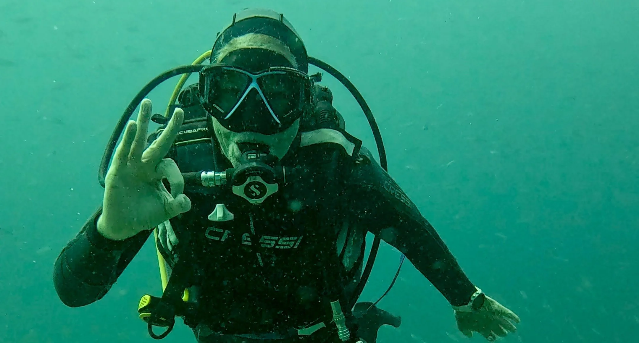 A scuba diver underwater making an OK gesture with her hand and wearing a black wetsuit, mask, snorkel, and scuba tank.