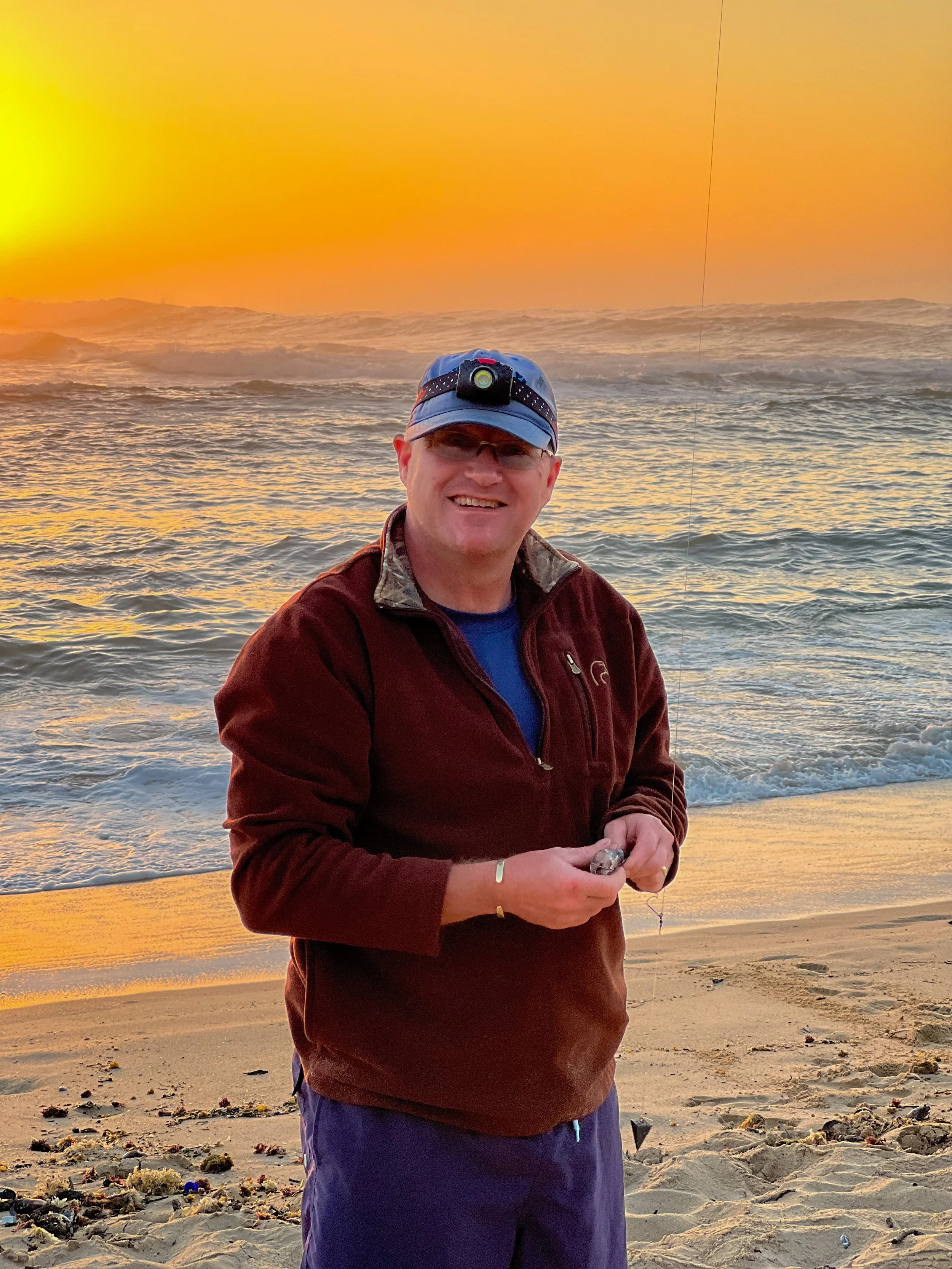 A man smiling at the camera on a beach at sunset, holding a small fish, with the ocean and waves behind him.
