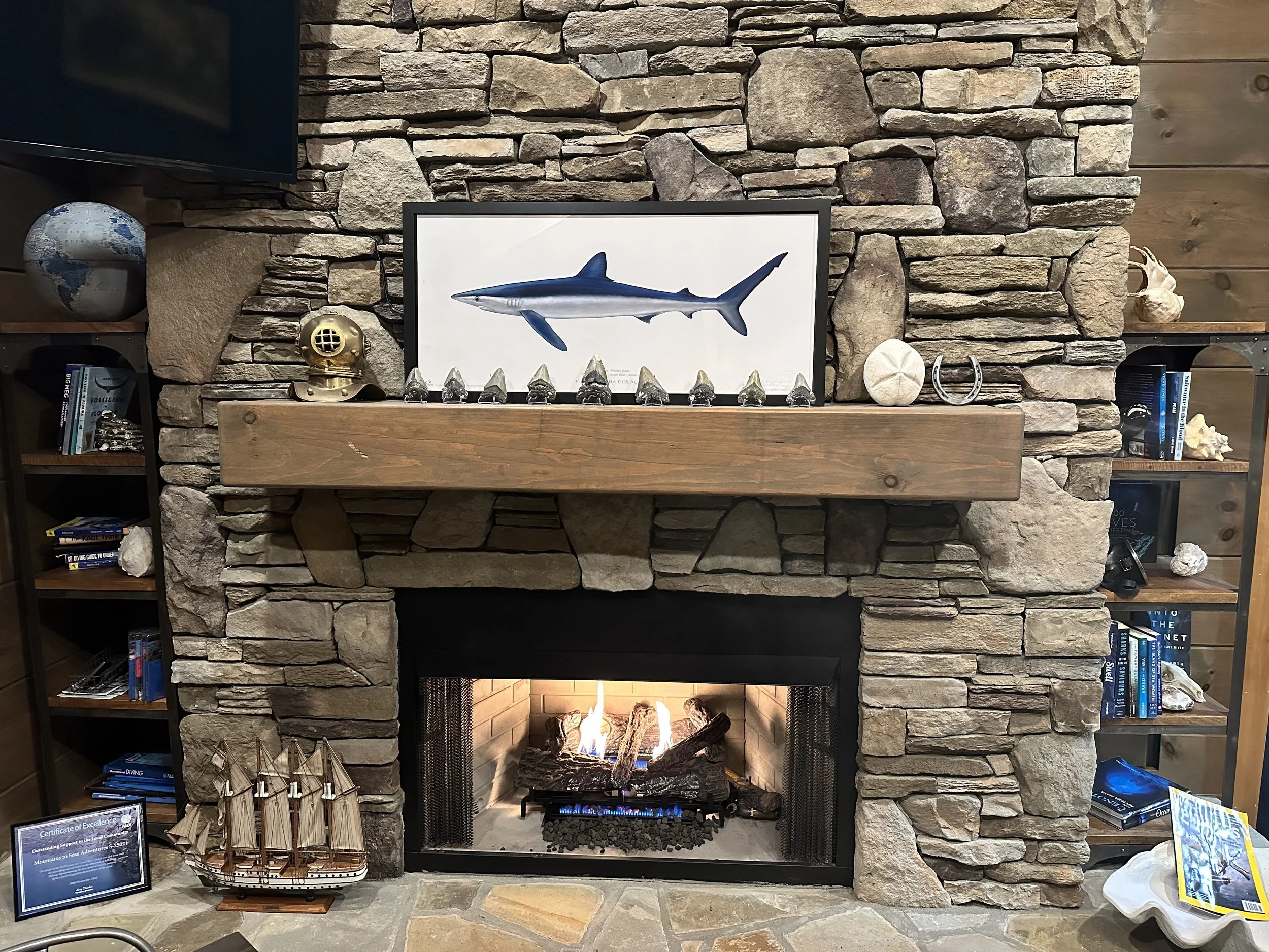 Stone fireplace with a wooden mantel, decorated with shells, a framed picture of a shark, a model ship, and various decorative objects, with a fire burning inside the fireplace.