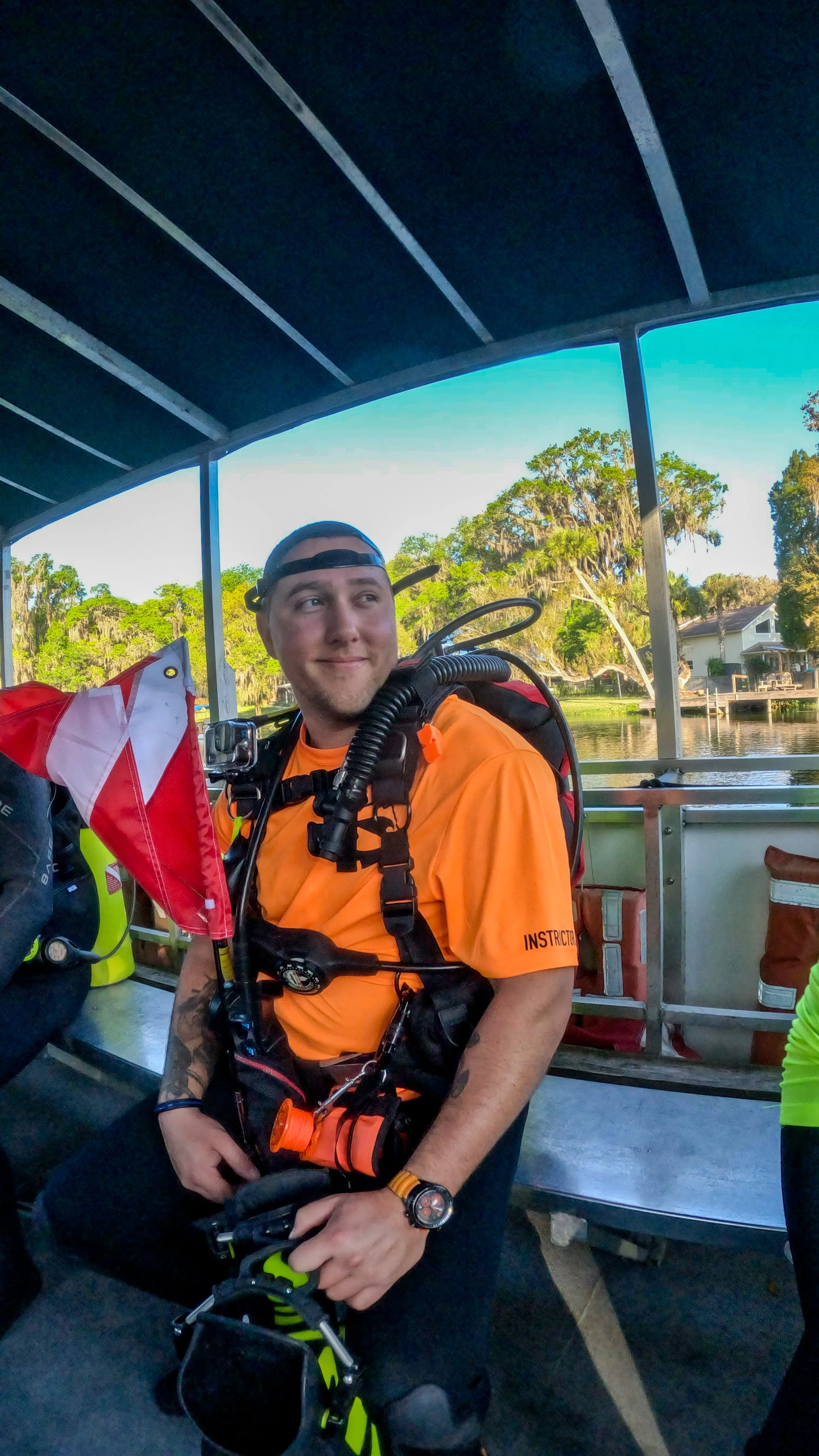 A young man in an orange shirt with a scuba mask on his head, standing on a boat with a body of water and trees in the background.