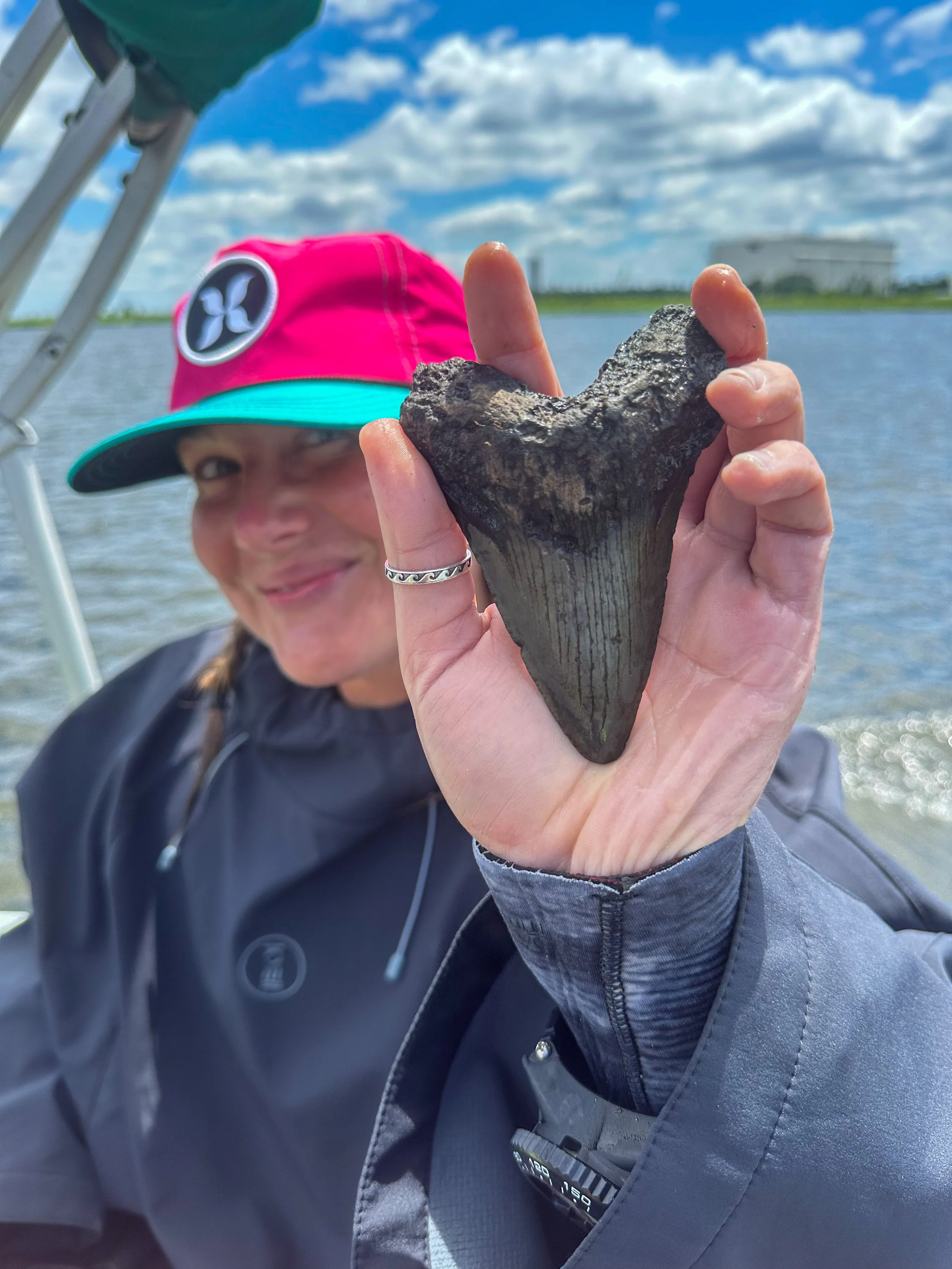 A person holding a large, dark gray shark tooth in front of their face, outdoors near a body of water with a cloudy sky.