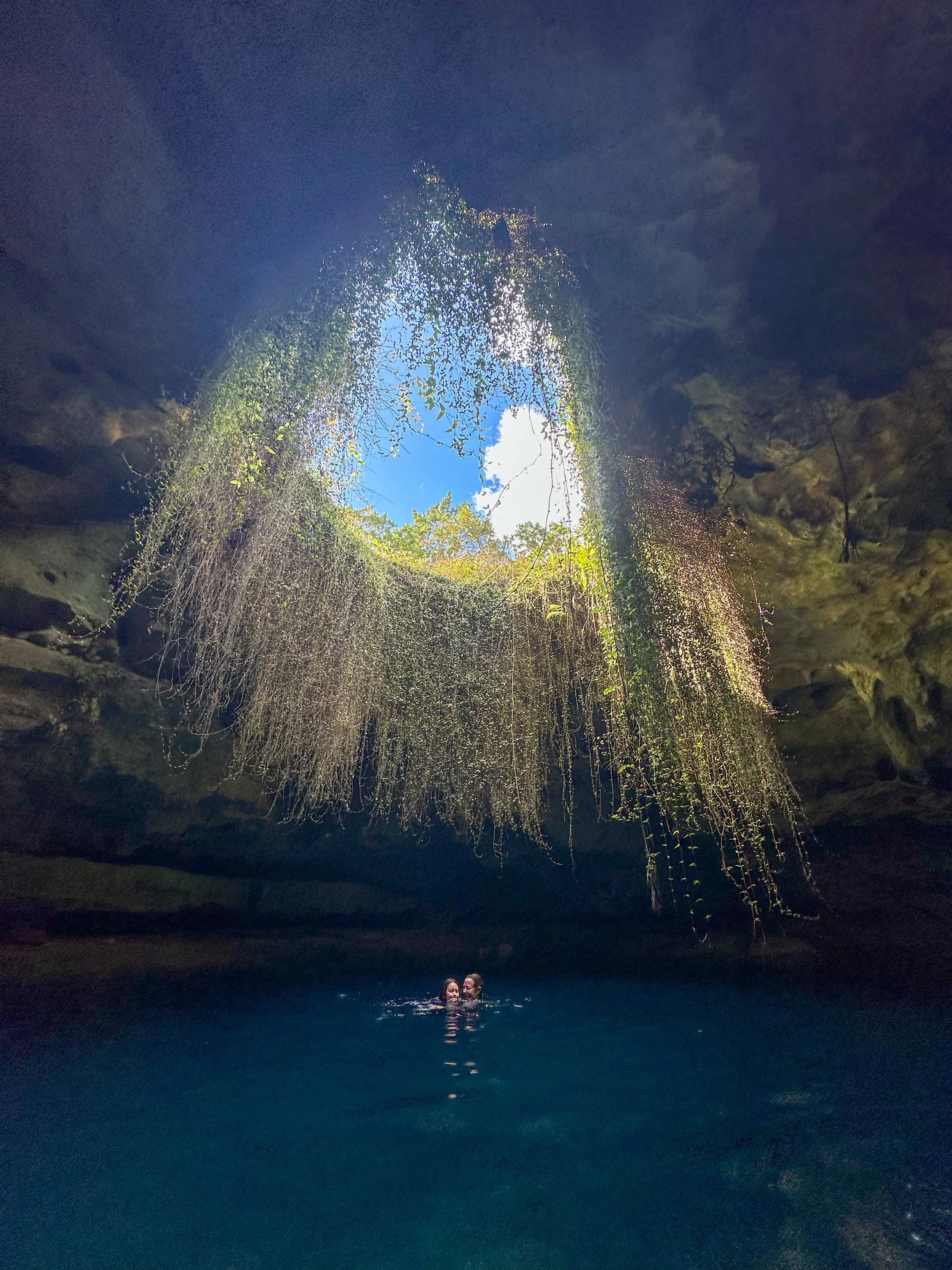 A cenote with a clear blue sky and clouds visible through an opening at the top. Two people are swimming in the water beneath the opening, surrounded by moss and hanging vines.