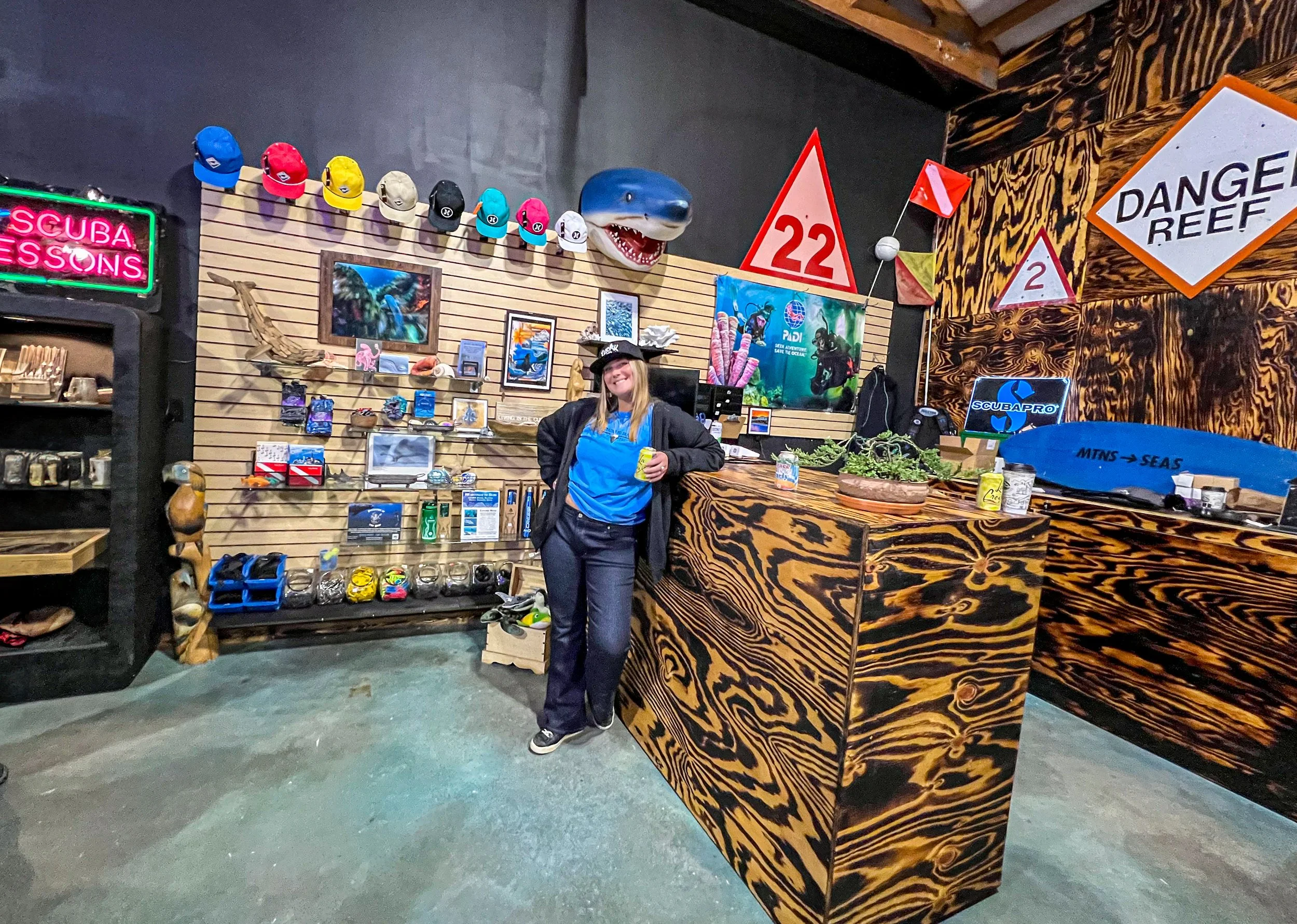 A woman standing behind a wooden checkout counter in a scuba diving shop. The background has a wall with hanging hats in various colors, framed pictures, and coastal decorations. There are scuba diving signs, a shark mask on the ceiling, and shelves 