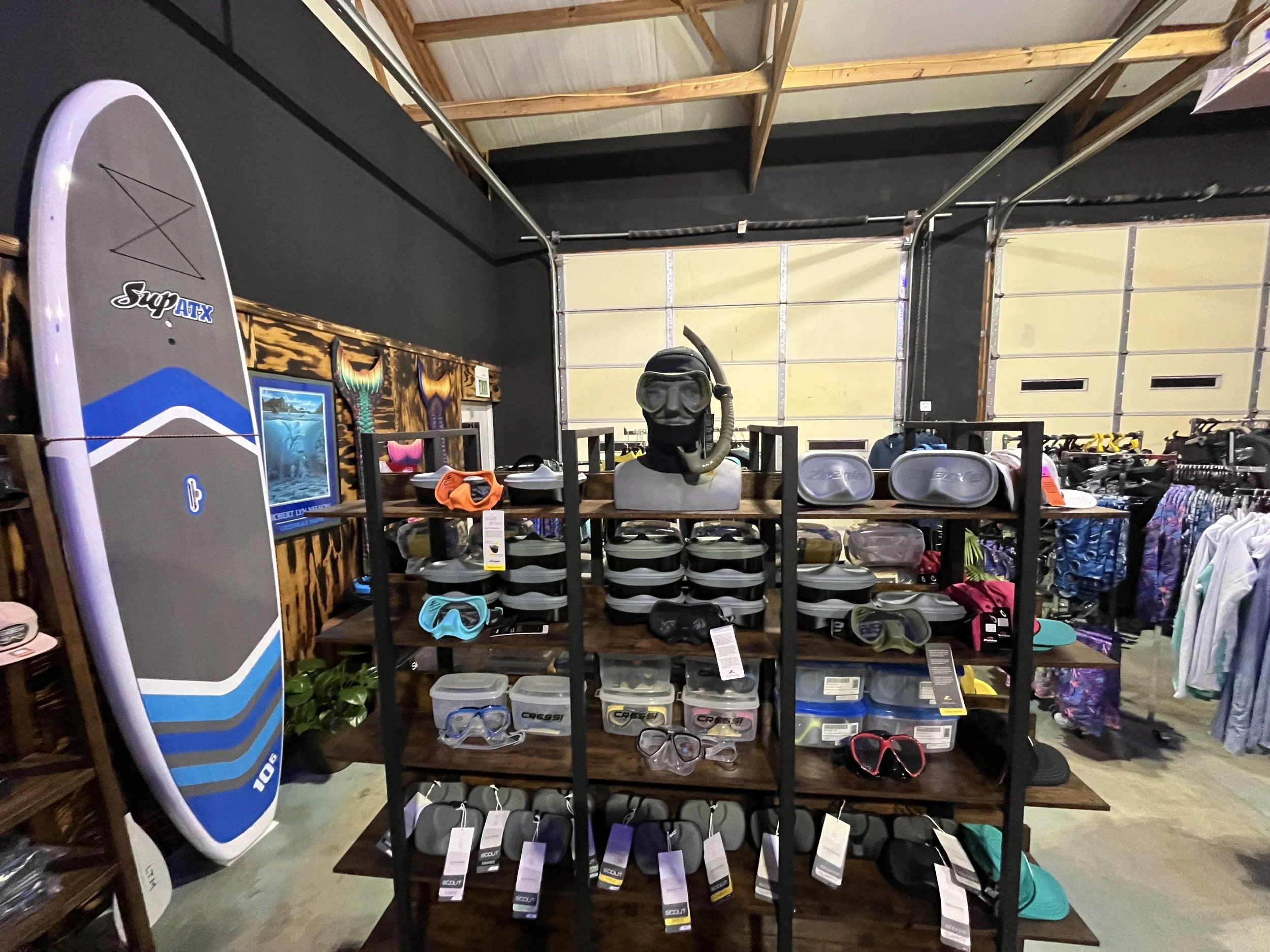 Shelves in a store with snorkeling masks, fins, and a scuba diving helmet, along with a stand-up paddleboard on the left side.