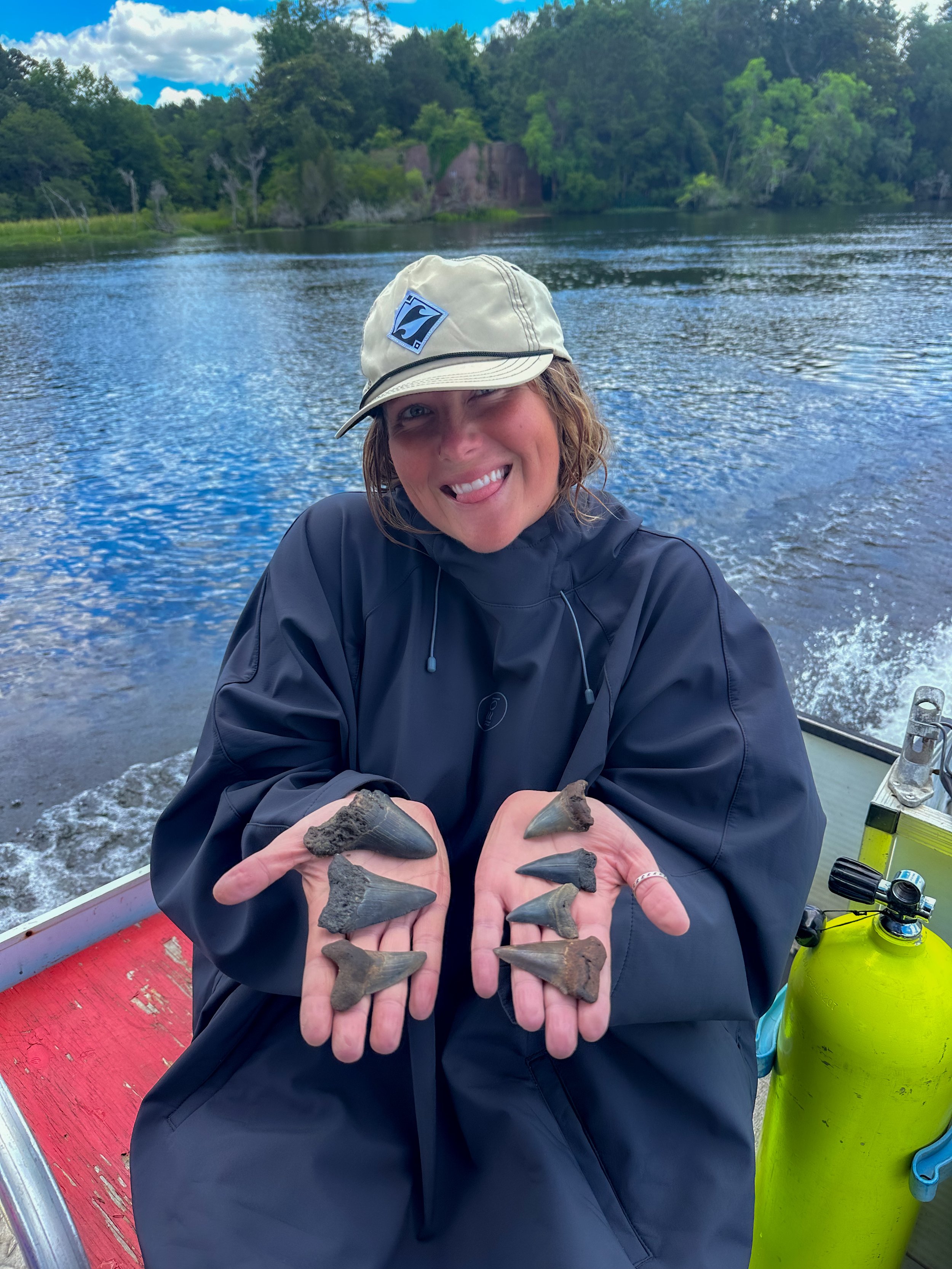 Woman on a boat holding several fossils of shark teeth, smiling with her tongue out, near a river with trees and blue sky in the background.