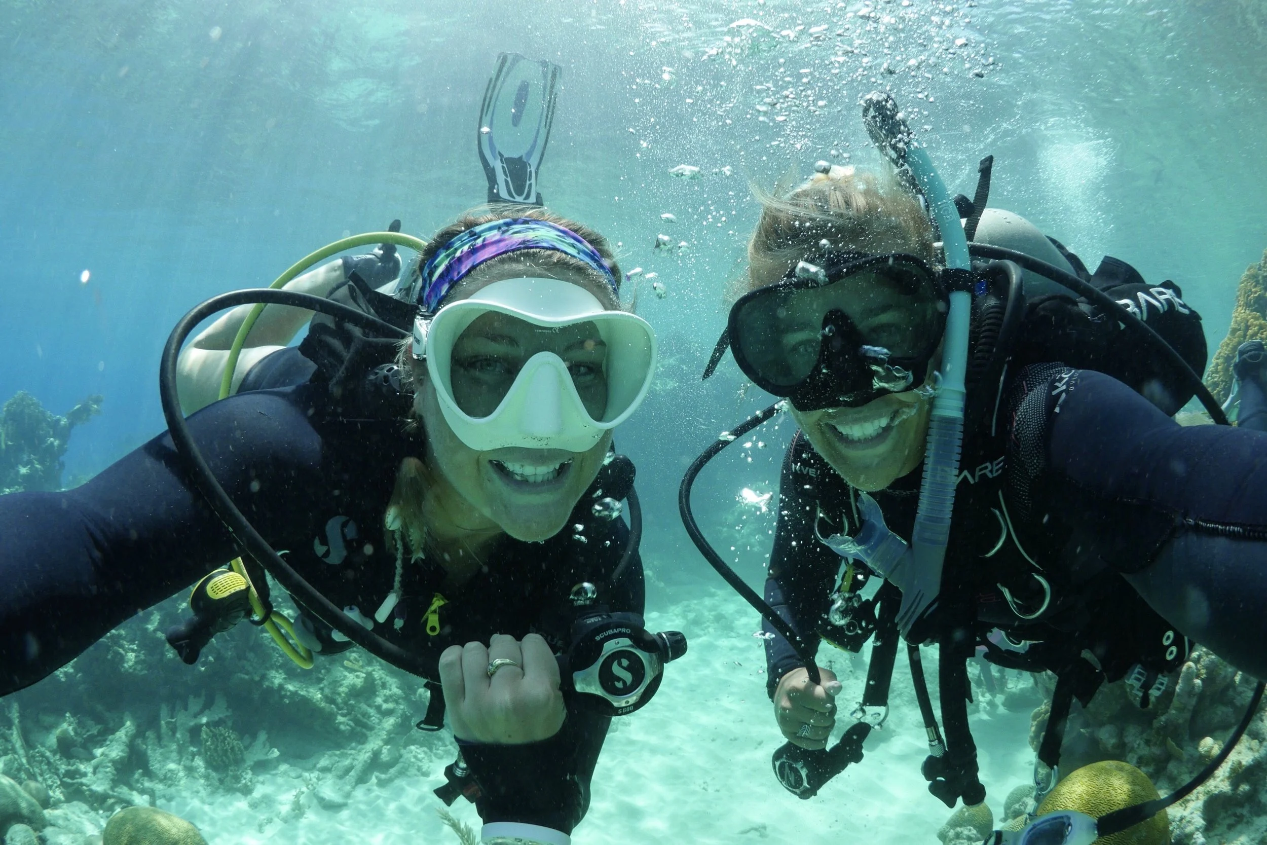 Two women scuba diving underwater, smiling for a selfie, with coral and fish in the background.