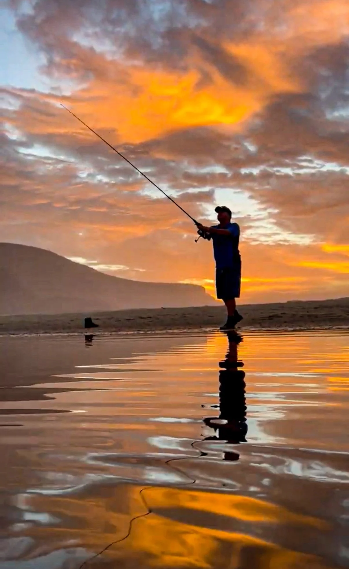 Person fishing on the beach at sunset with orange and purple clouds reflected on the wet sand.