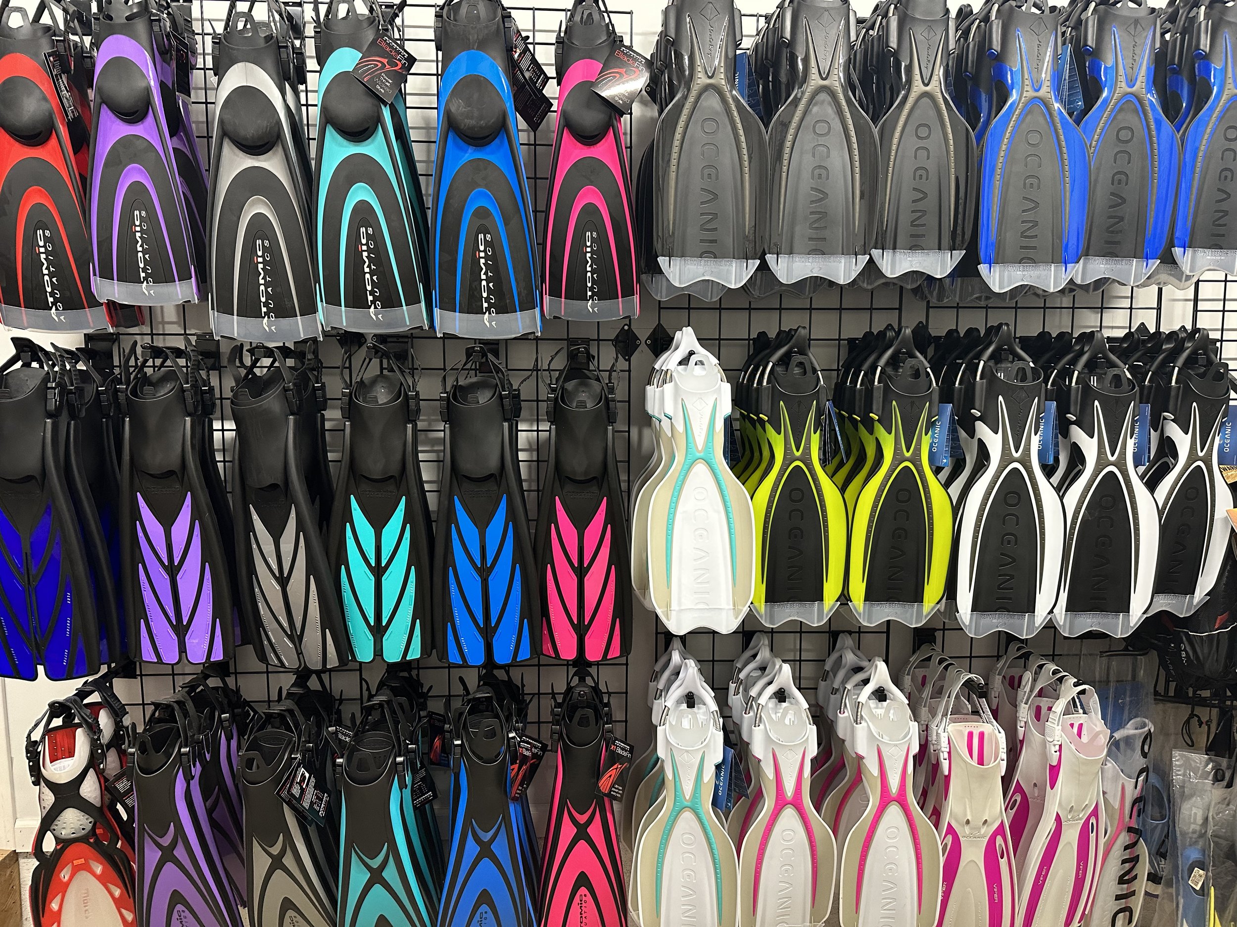 A display of various colored snorkeling fins hanging on a grid wall in a store, including black, purple, gray, blue, pink, white, yellow, and orange fins.