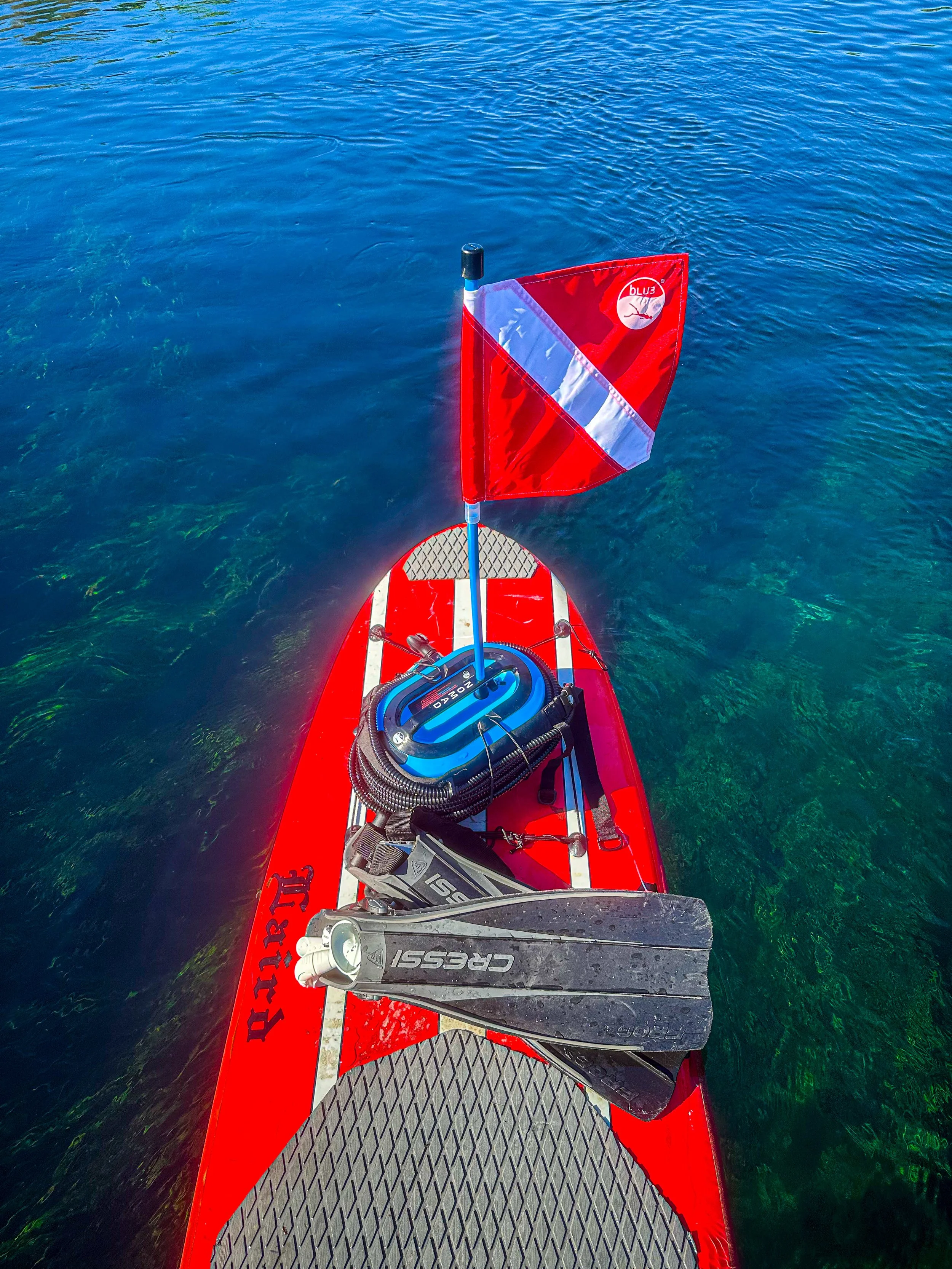 Red paddleboard with a red and white flag, paddle, diving fins, and waterproof gear on calm blue water.