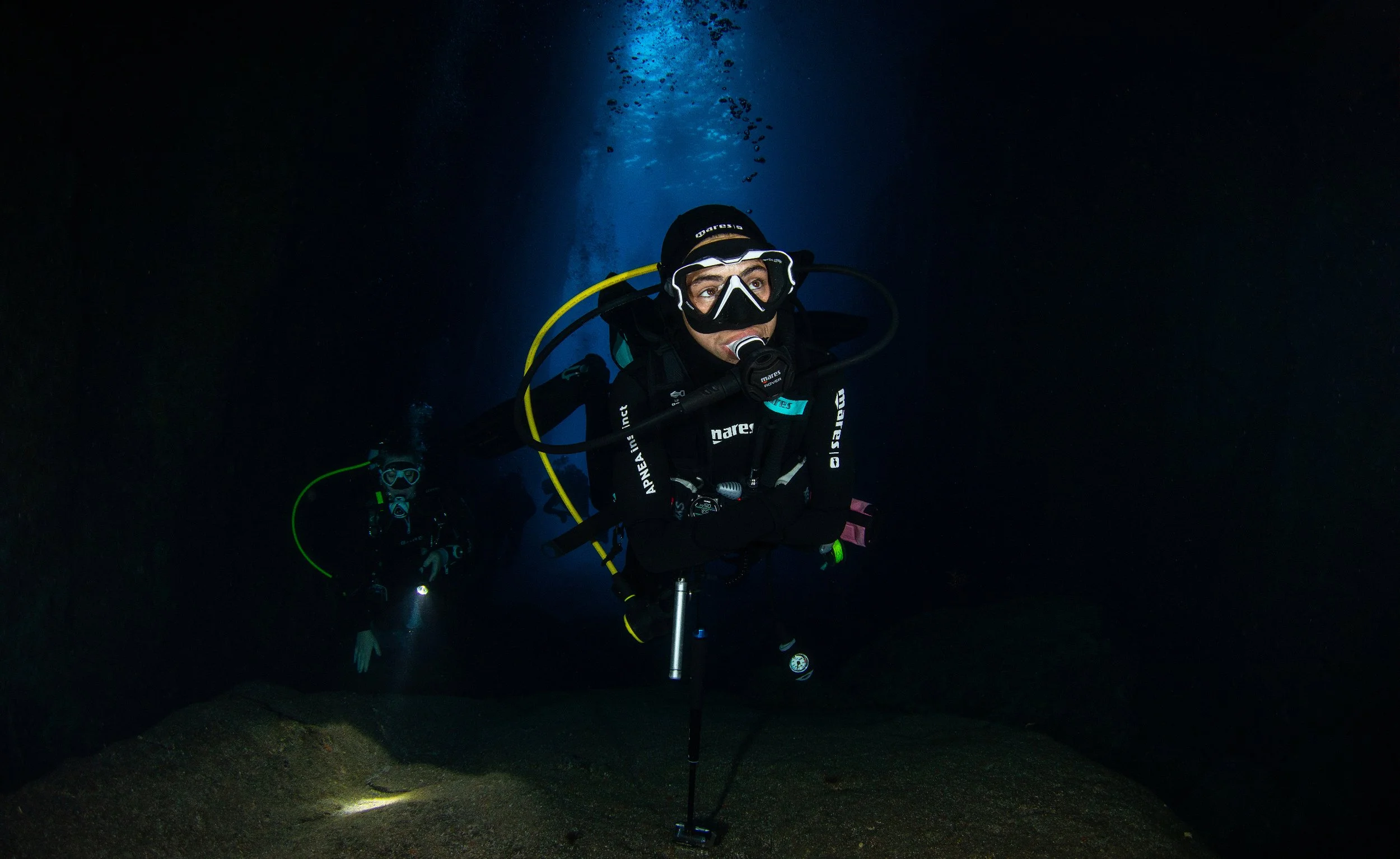 Two scuba divers exploring underwater cave, with one diver in foreground and another in background, illuminated by flashlights.