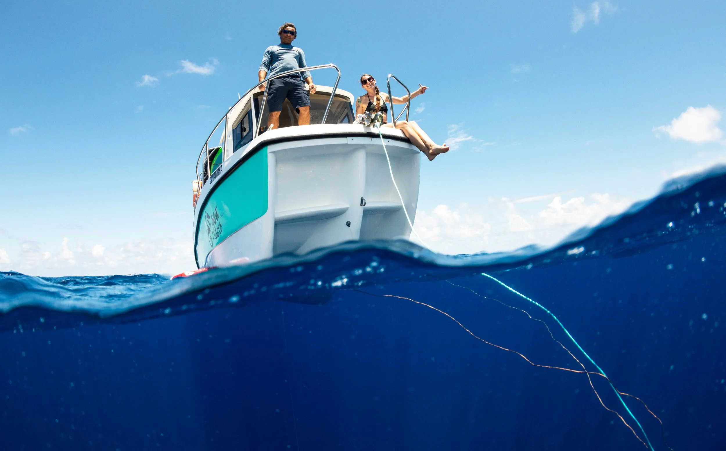 People on a boat with one woman sitting on the edge, fishing or relaxing, and a man standing nearby, against a blue sky with scattered clouds.