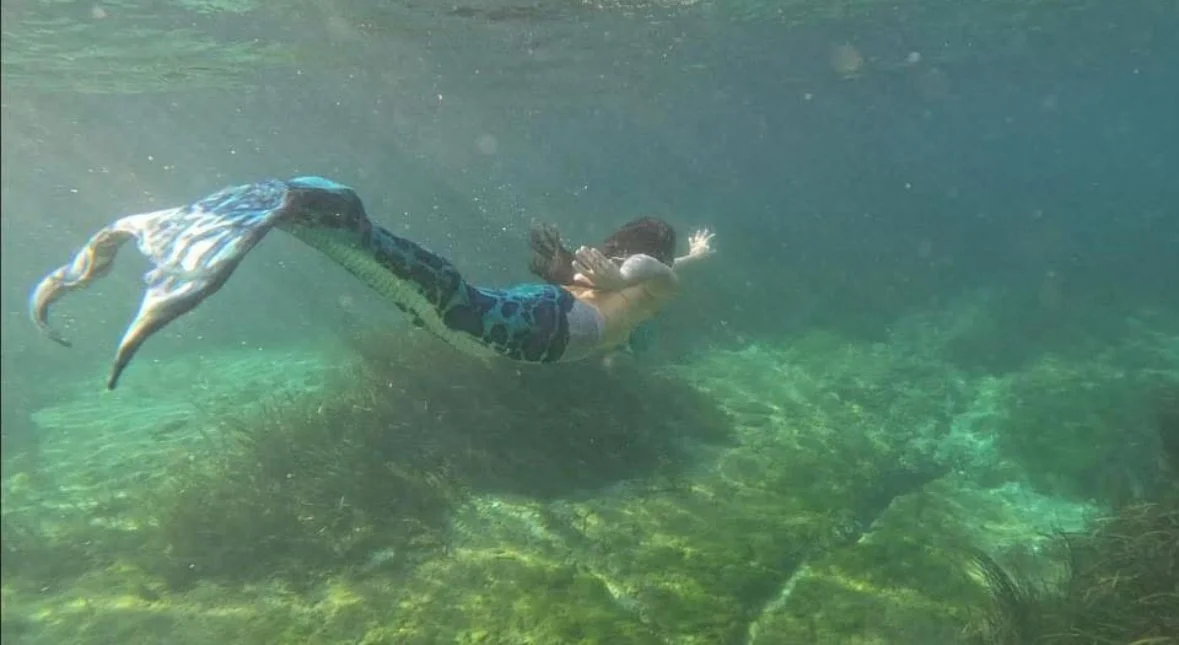 Child swimming underwater in a lake or pond, wearing blue swim trunks, with green aquatic plants and algae on the bottom