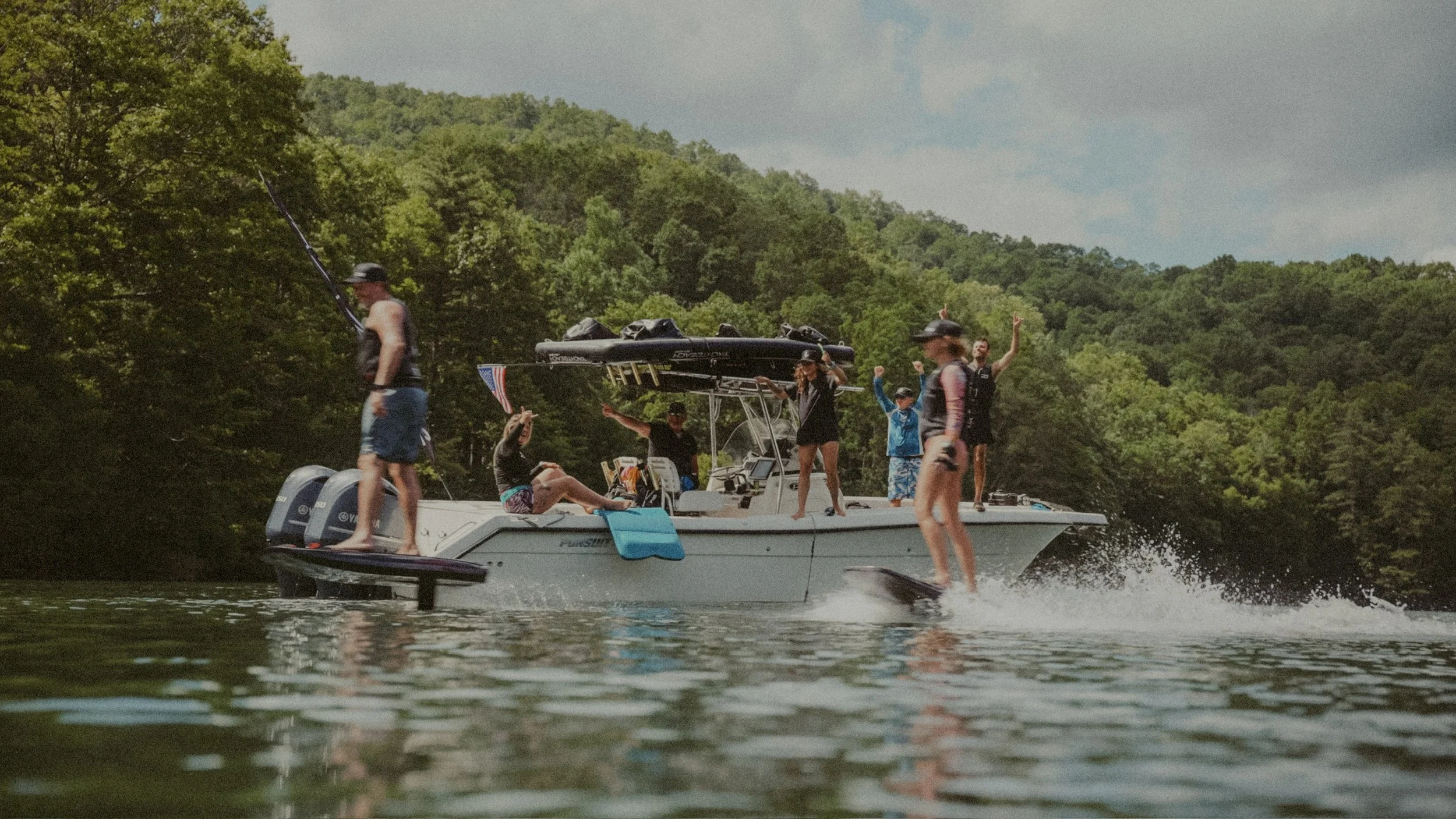 A group of people enjoying water activities on a boat and on paddleboards on a lake surrounded by green trees and hills.