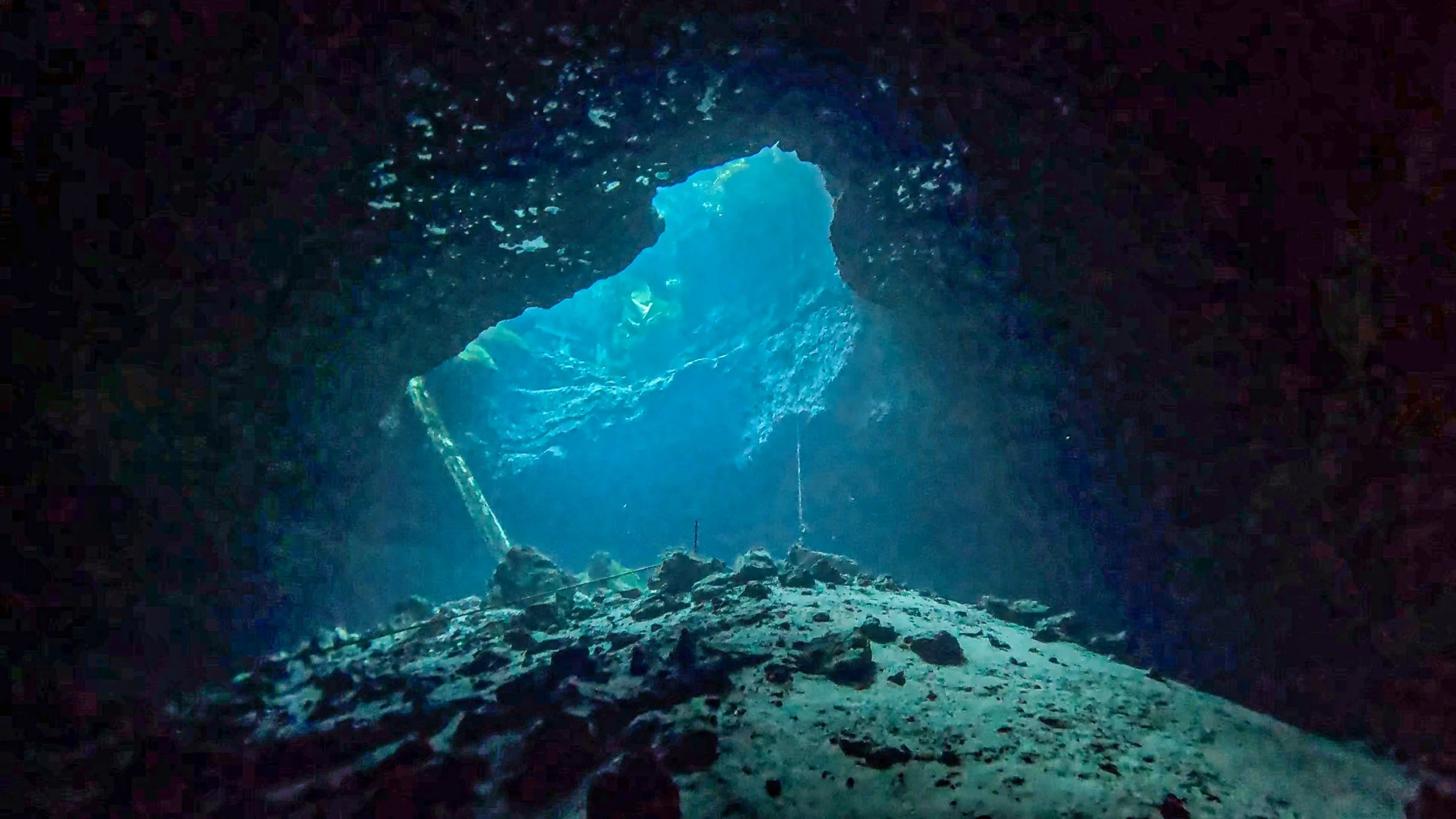 Underwater cave with sunlight shining through the opening, rocky floor, and walls covered in dark textures.