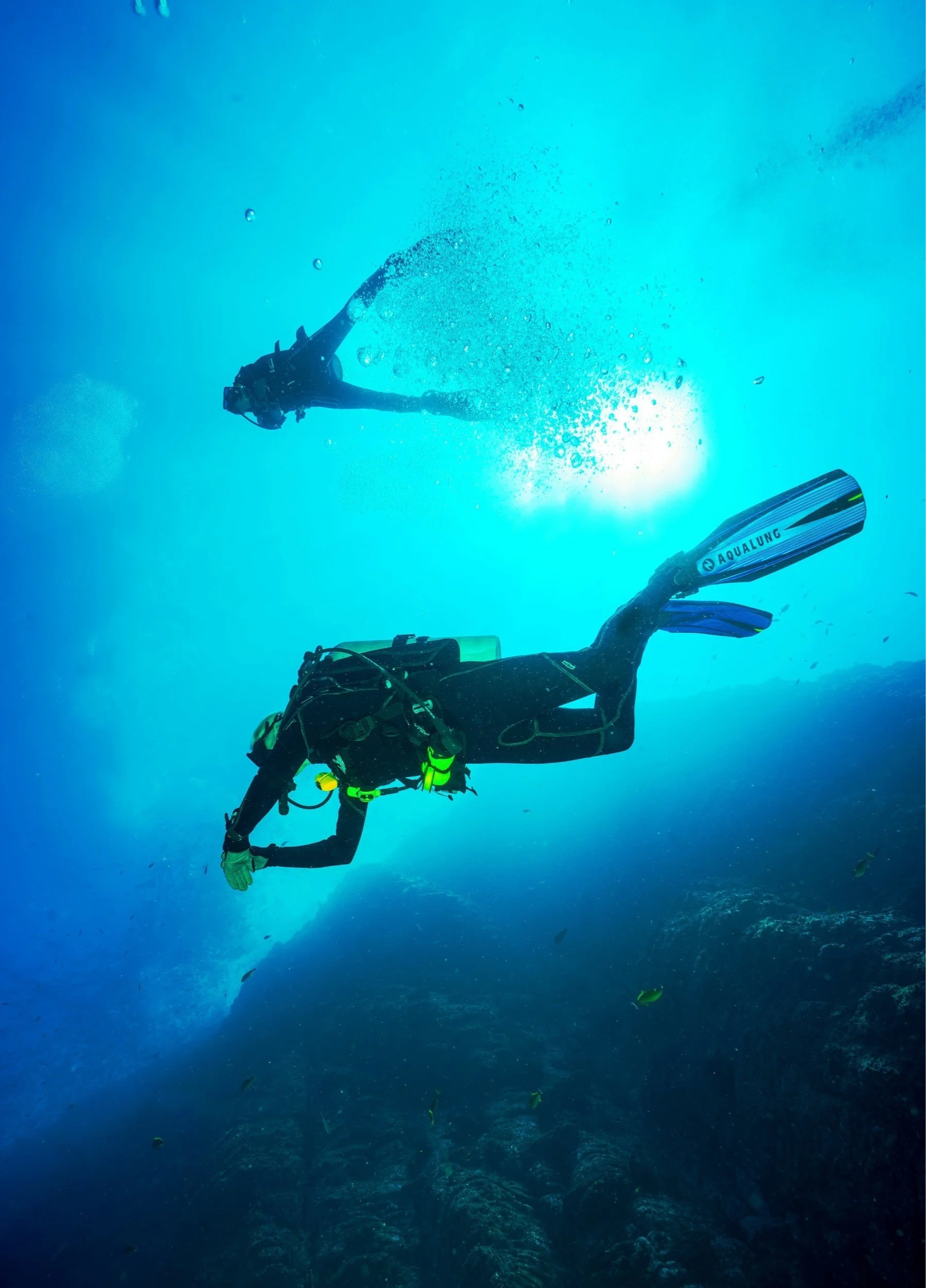 A scuba diver in black gear swimming underwater near rocks with another diver in the background.