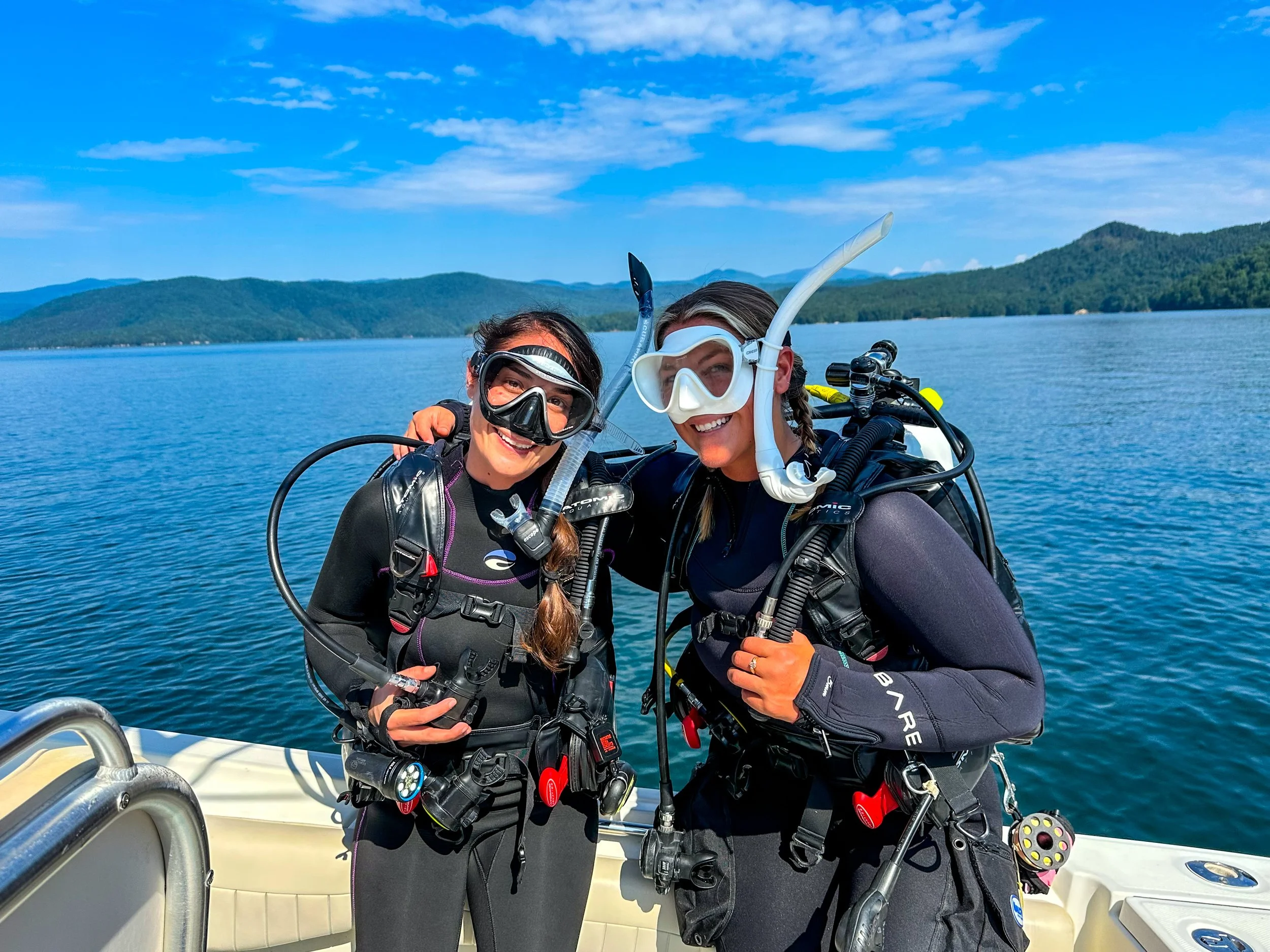 Two women in scuba diving gear on a boat, smiling, with a lake and mountainous landscape in the background.