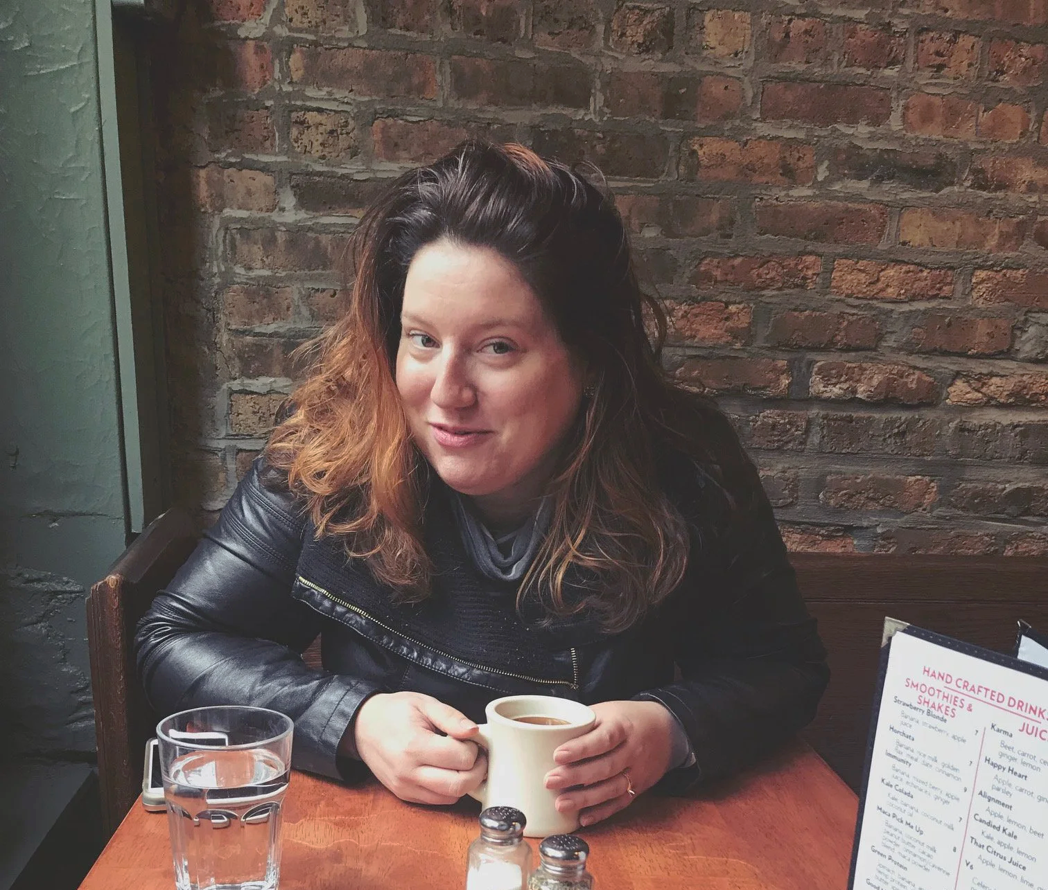 A woman in a faux leather jacket is drinking coffee. Her head tilts to the left and she makes eye contact with the camera.