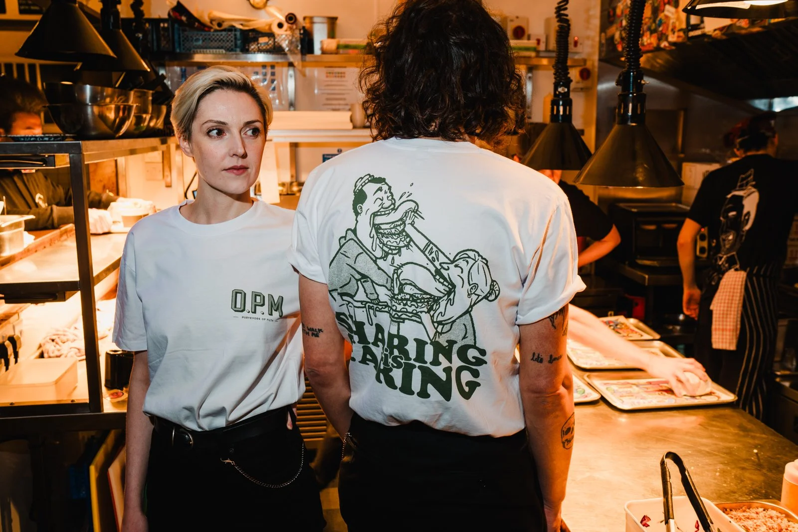 Photograph of two people stood in a kitchen wearing matching white t-shirts featuring green illustrations of a couple stuffing cartoonishly large burgers into each other's mouths. Underneath it says 'Sharing is caring' in a bold font.