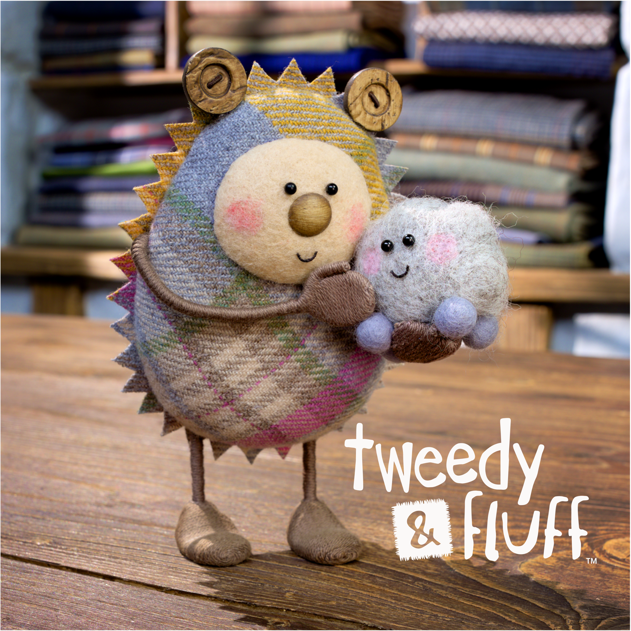Promotional image of Tweedy and Fluff. Two handmade stop motion puppets, Tweedy is made of a checked tweed resembling a mixture between a hedgehog and koala, holding fluff who resembles a dust bunny or ball of dryer lint with a face and four legs.