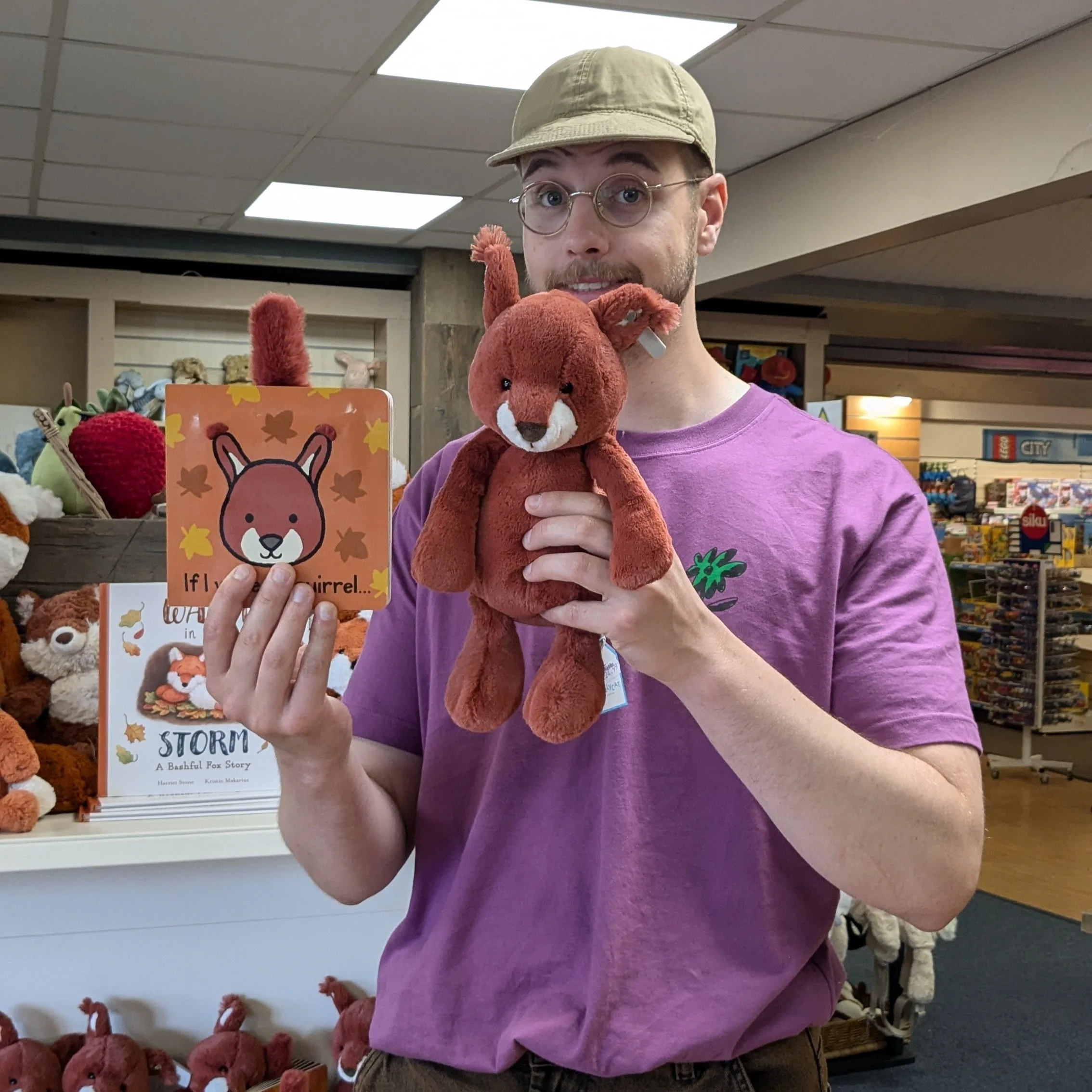 Image of a man standing in a shop, holding a picture book of squirrel in his right hand and an accompanying plush toy of the squirrel in his left. He is smiling proudly at the camera.