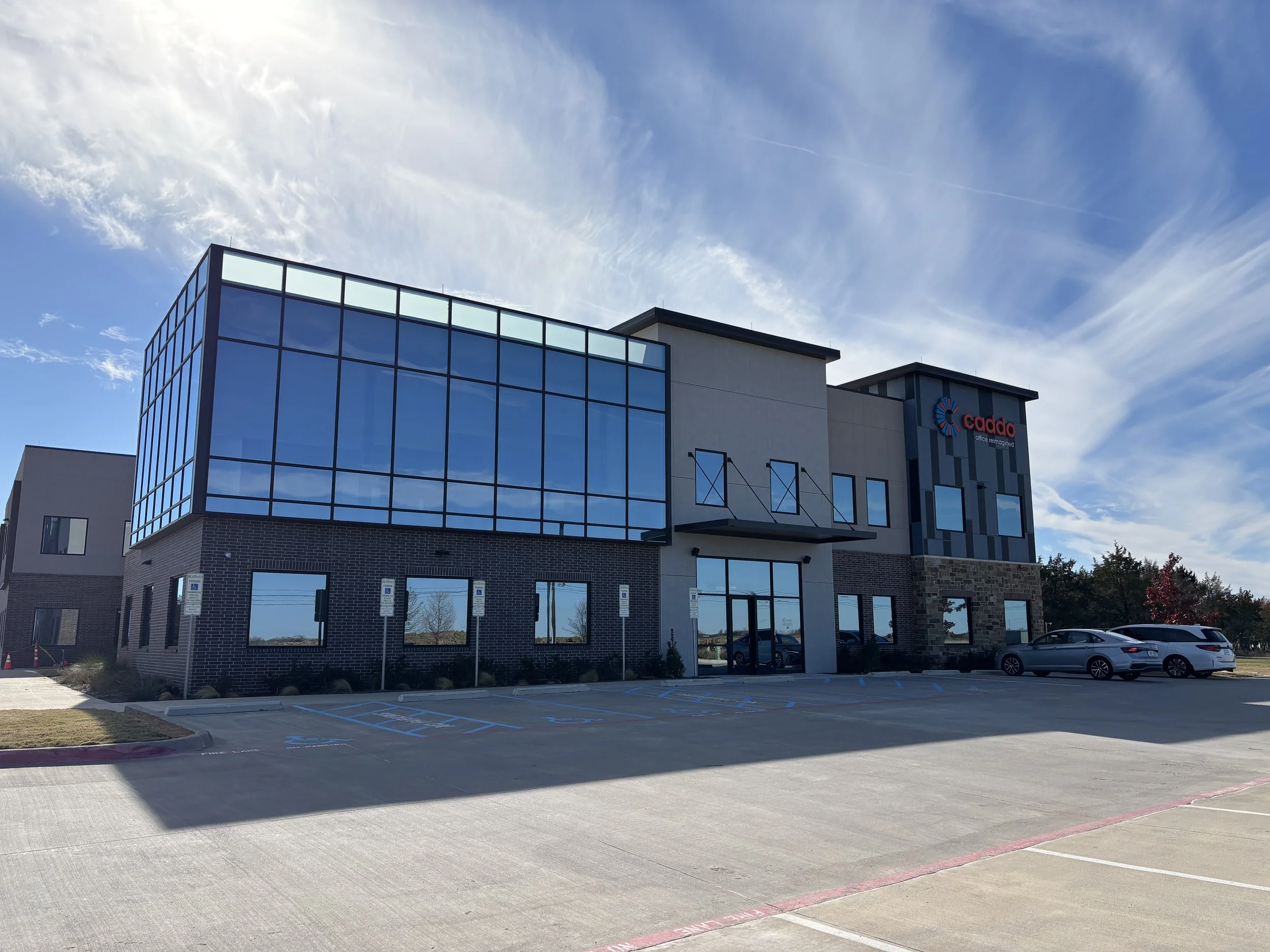 Modern office building with a glass facade, parking lot with several cars, and a blue sky with clouds. The building has the Caddo logo.
