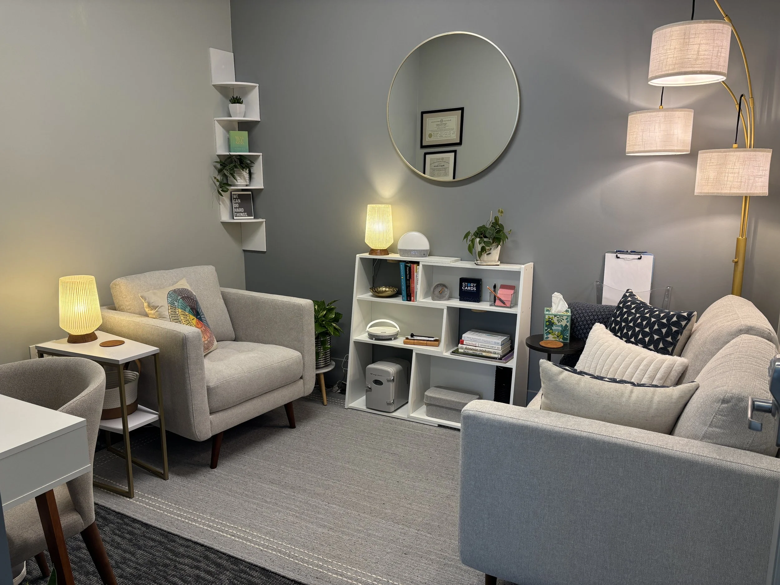Comfortable therapy room with gray walls and furniture, including couches, side tables with lamps, and a white shelving unit with decorative items and books. Wall decor includes a round mirror and framed certificates.