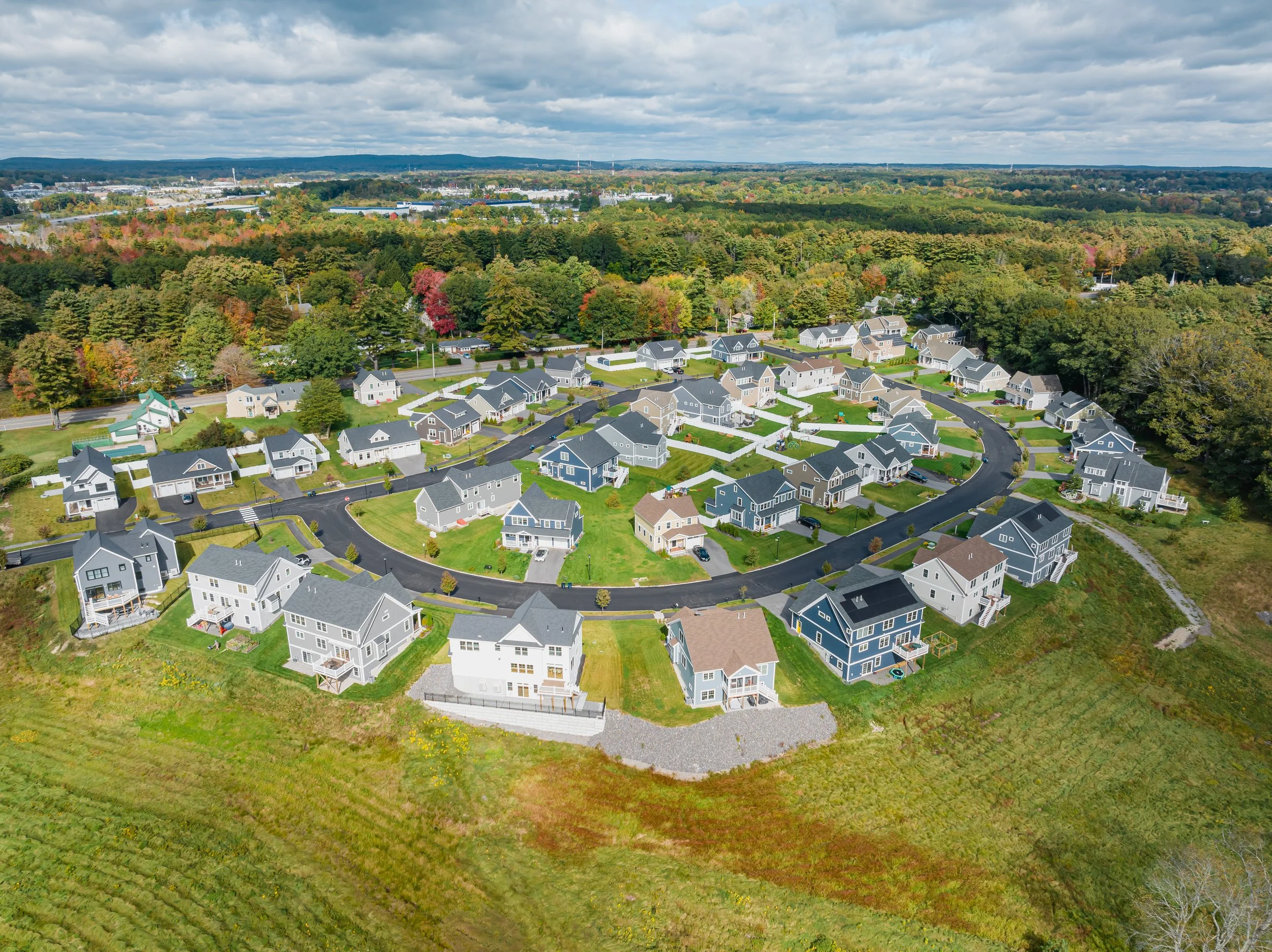 An aerial view of a suburban neighborhood with multiple houses arranged in a circle, surrounded by green lawns, trees, and a wooded area in the background under a partly cloudy sky.