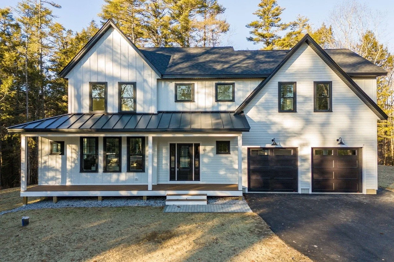 Front view of a modern two-story house with white siding, black window frames, and black garage doors, surrounded by trees and a clear sky.