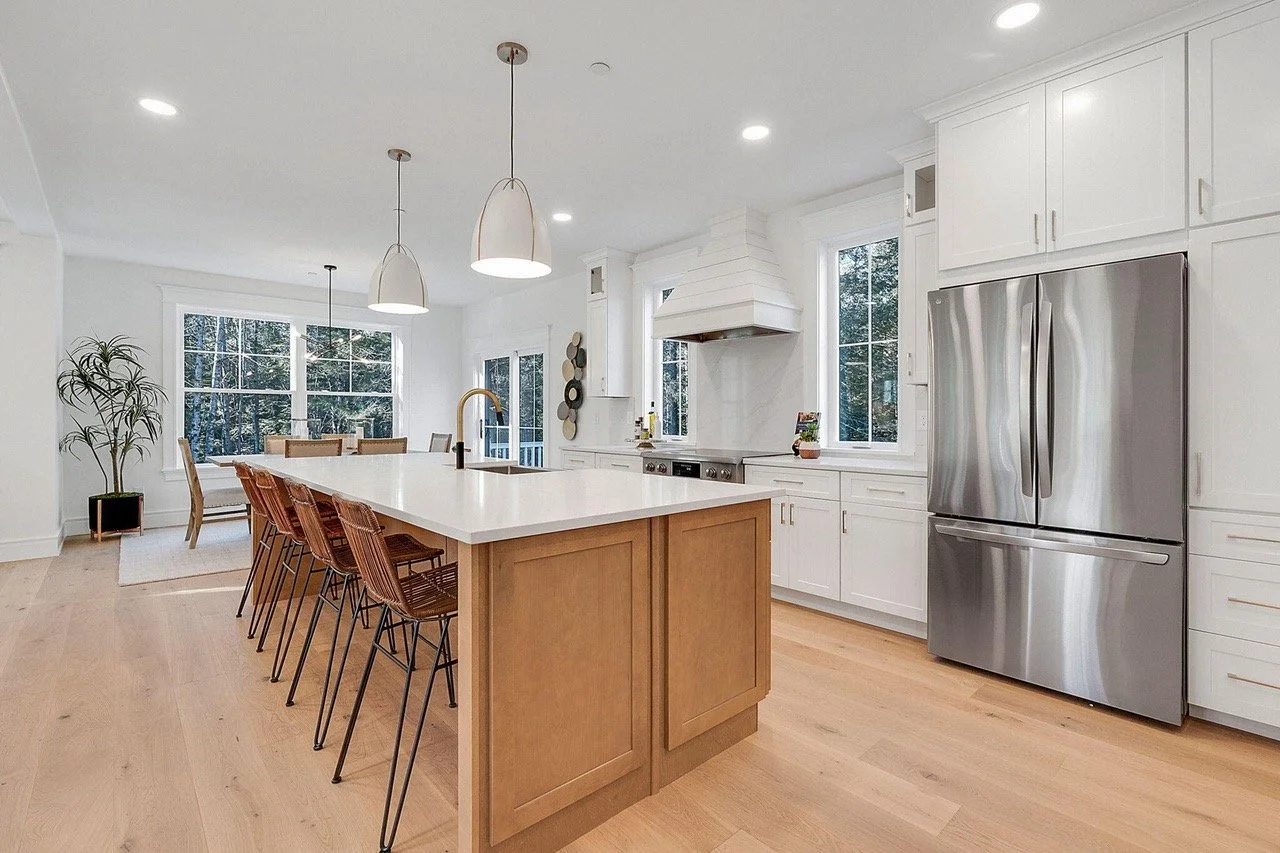 Modern kitchen with white cabinets, stainless steel refrigerator, wooden kitchen island, and large windows with a view of trees.