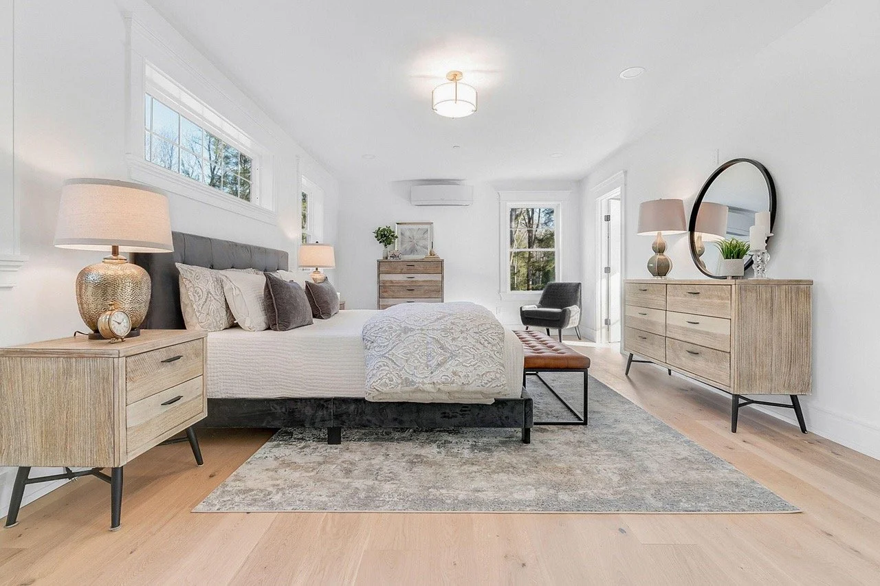 Bright bedroom with a large bed, nightstands with lamps, a dresser, an armchair, and a window showing trees outside.