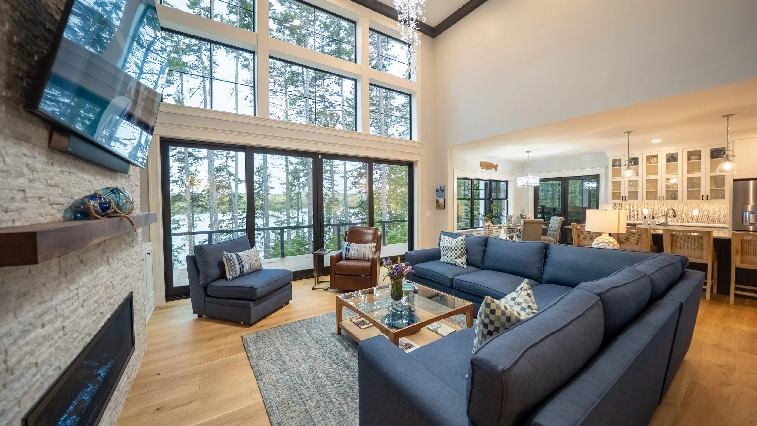 Living room with large windows showing trees outside, a blue sectional sofa, a blue armchair, a wooden coffee table with flowers, and a TV on the wall. Kitchen with island and bar stools visible in the background.